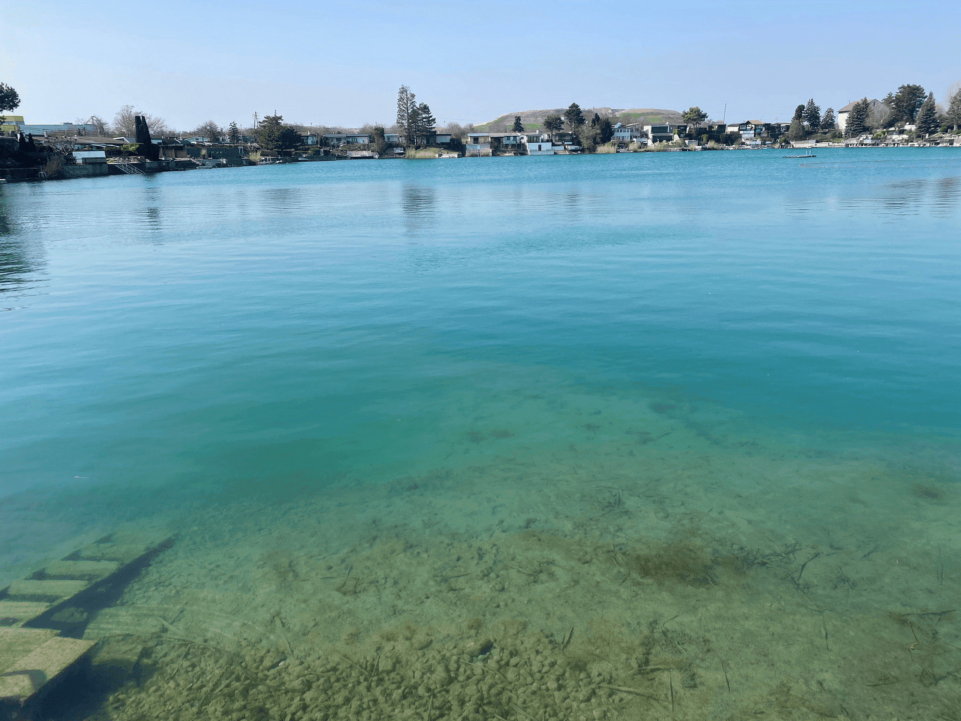 Klares, türkisfarbenes Seewasser mit sichtbarem Grund und Häusern am gegenüberliegenden Ufer.