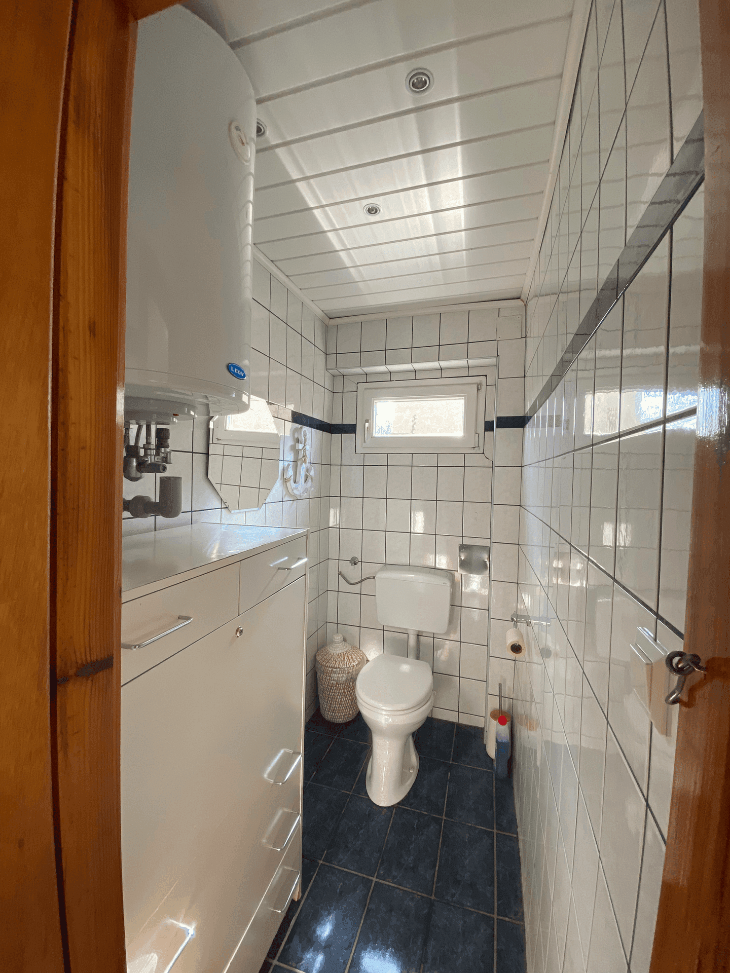 Kleines WC mit Toilette, Boiler und weiß-schwarzen Fliesen, gut beleuchtet.