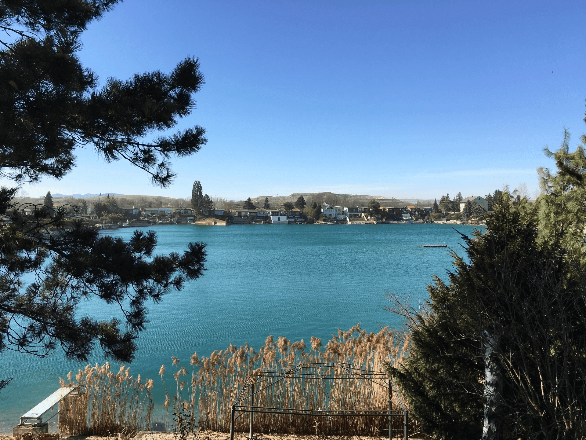 Weitläufiger Blick über den See mit Häusern am gegenüberliegenden Ufer unter blauem Himmel.