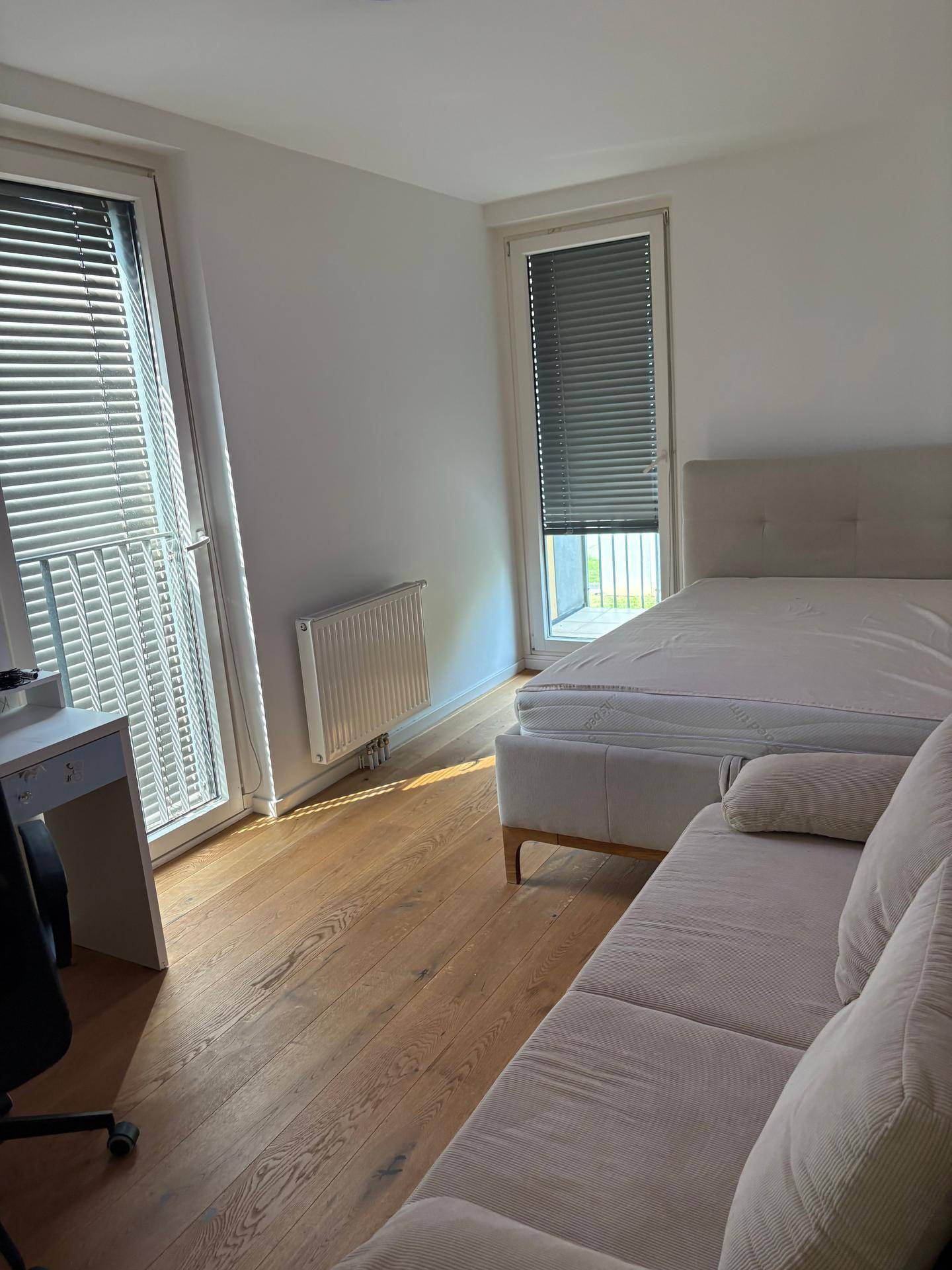 Helles Schlafzimmer mit Bett, Holzboden und großen Fenstern mit Jalousien.