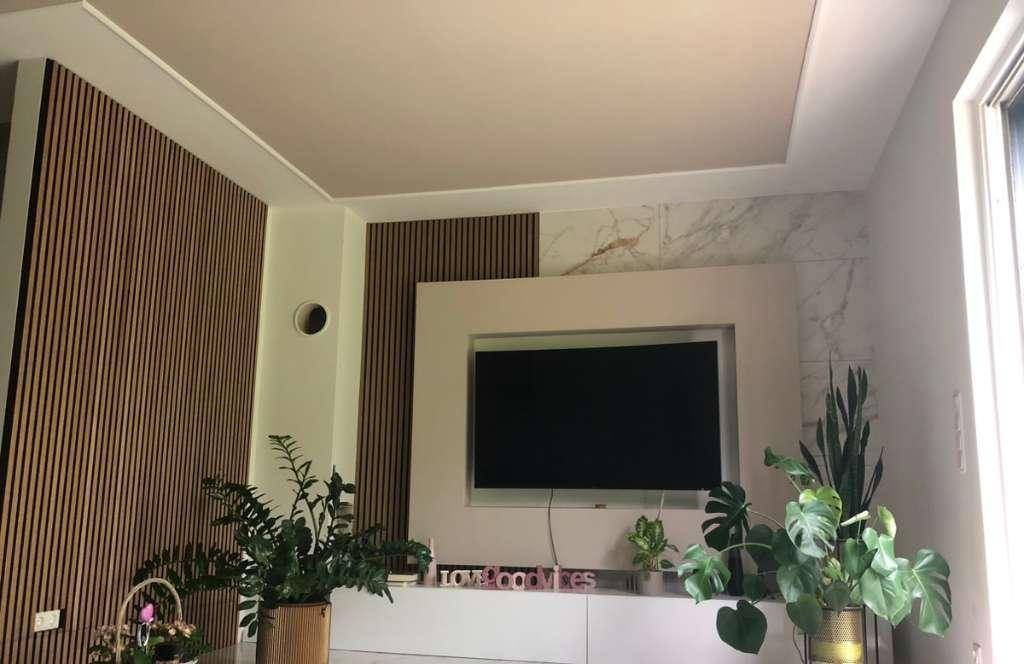 Modernes Wohnzimmer mit TV-Wand, Holzlamellen und indirekter Deckenbeleuchtung, dekoriert mit Pflanzen.