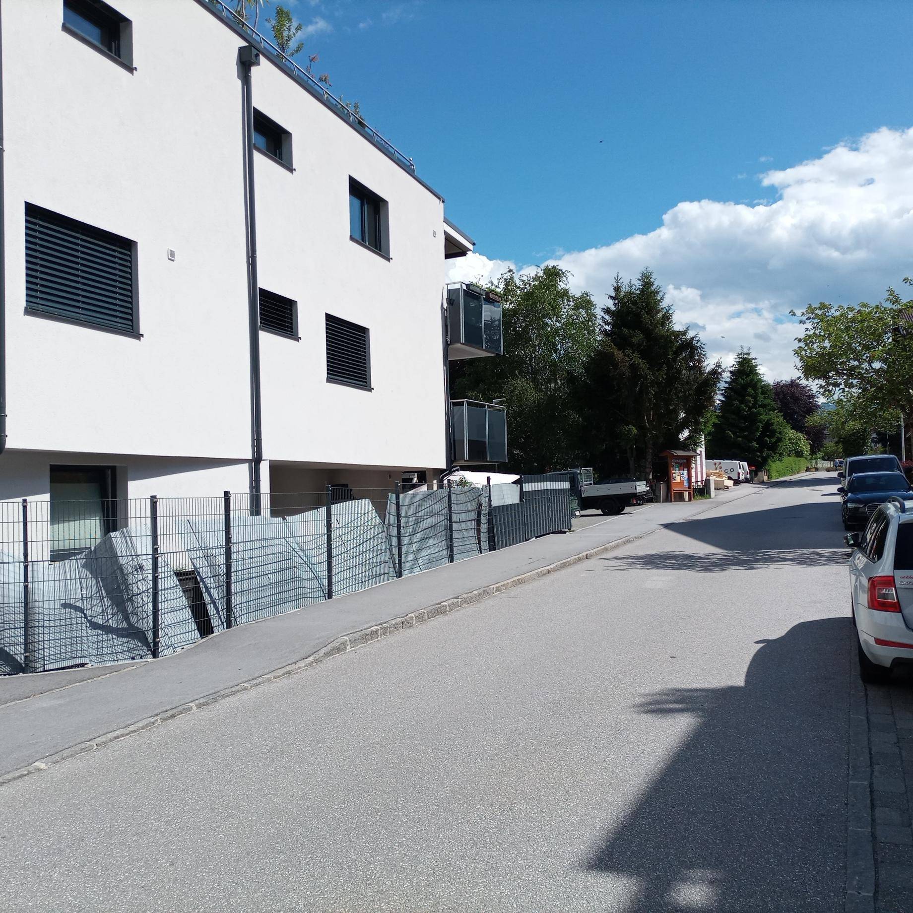 Modernes weißes Gebäude mit dunklen Fensterläden entlang einer Straße mit parkenden Autos und grüner Umgebung.