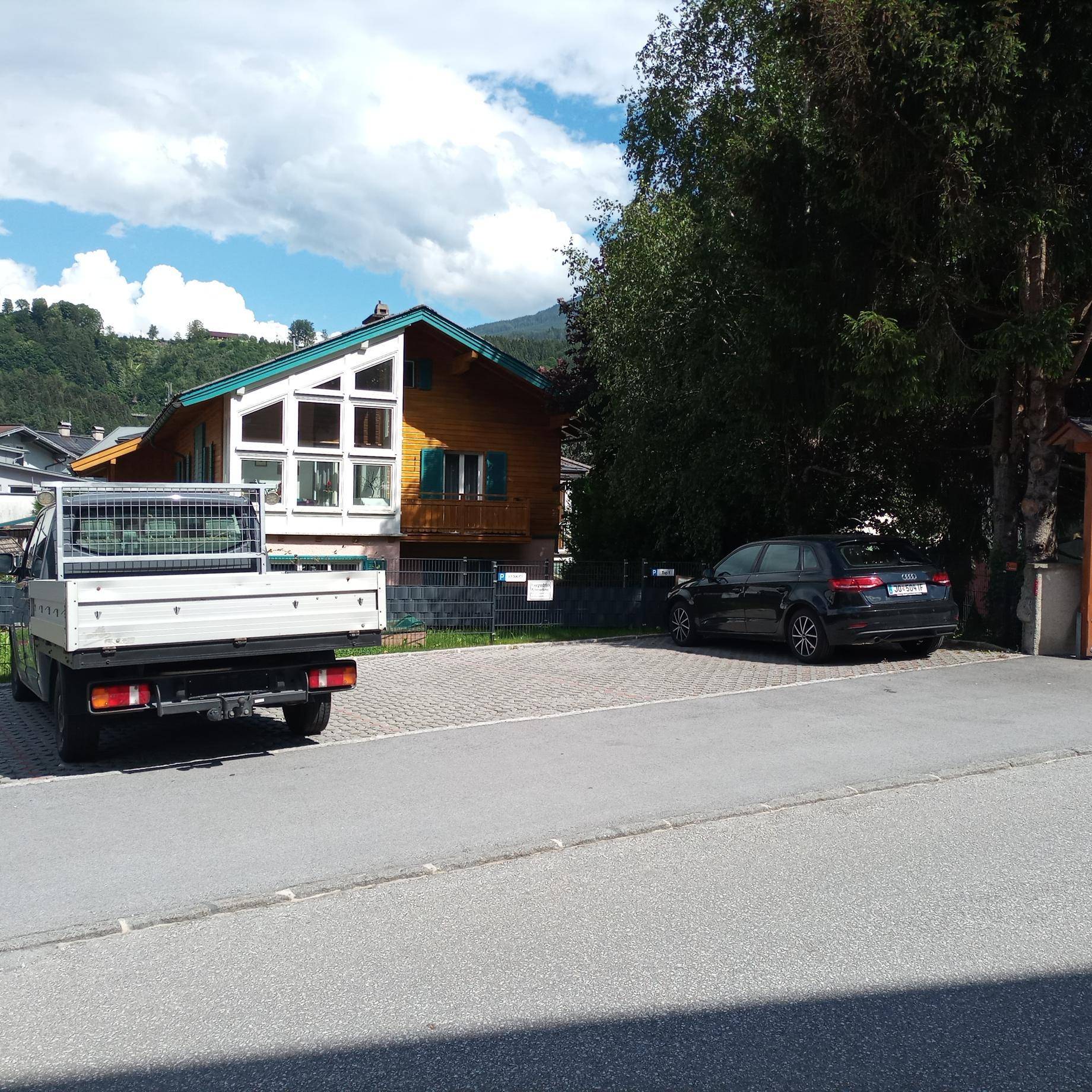 Holzhaus im alpinen Stil mit großen Fenstern und Parkplätzen davor, umgeben von grüner Landschaft.
