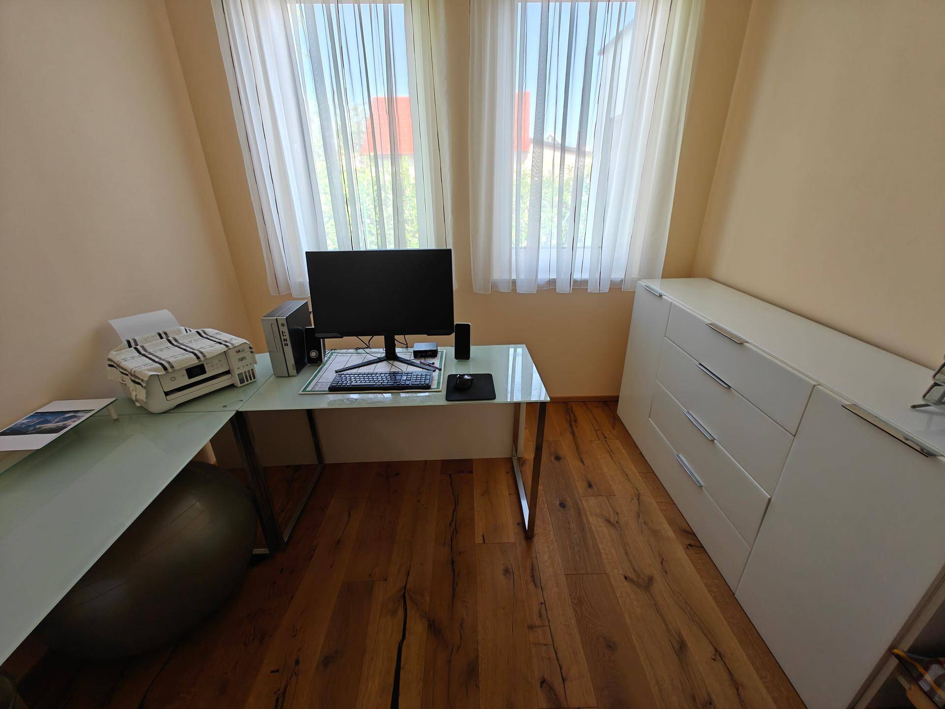 Arbeitszimmer mit modernem Schreibtisch und weißer Kommode unter den großen Fenstern.