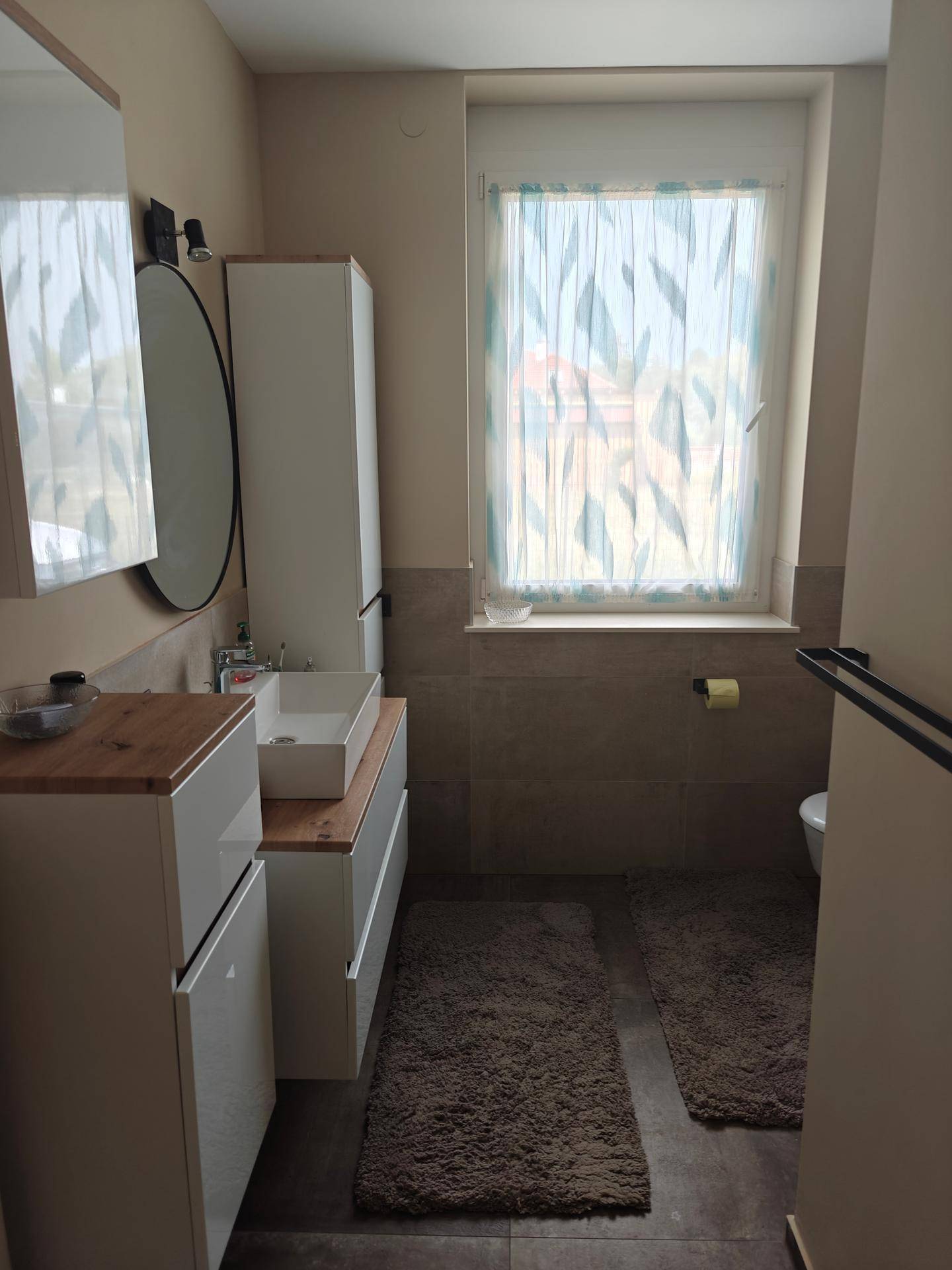 Modernes Badezimmer mit Doppelwaschbecken, großem Spiegel und einem Fenster mit heller Gardine.