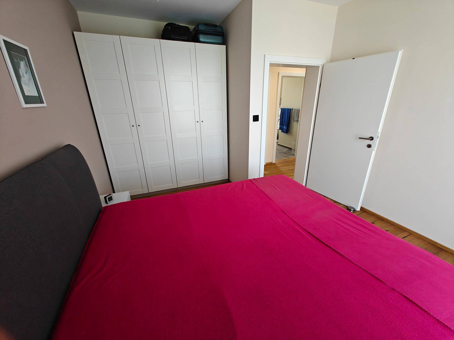 Schlafzimmer mit Bett und weißem Kleiderschrank, mit Zugang zu einem weiteren Raum.