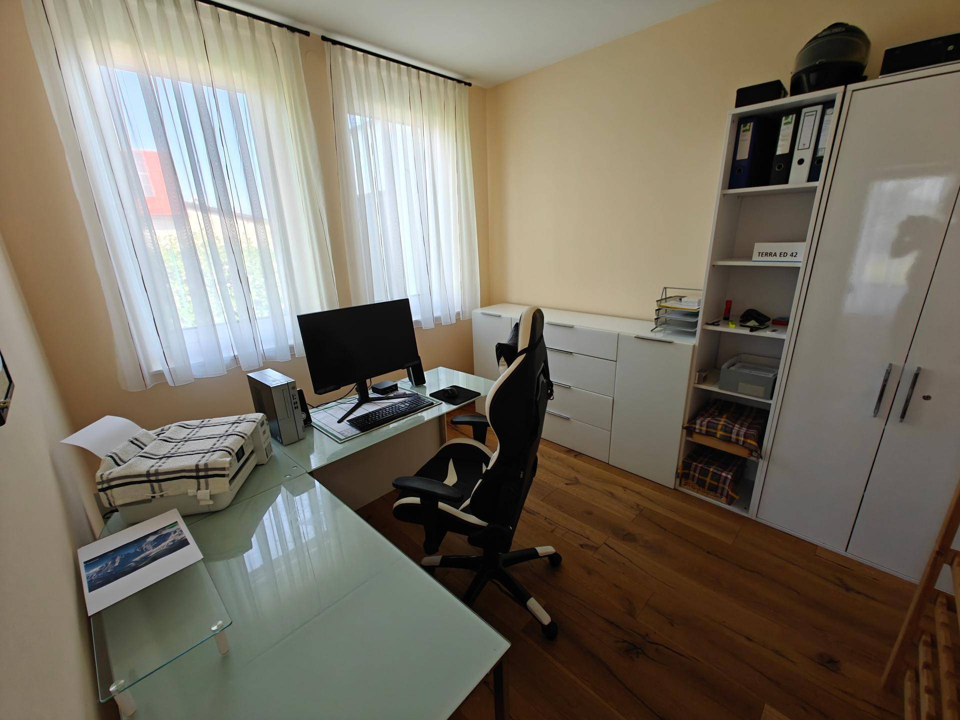 Helles Arbeitszimmer mit Schreibtisch, Computer und weißen Regalen und Schränken.