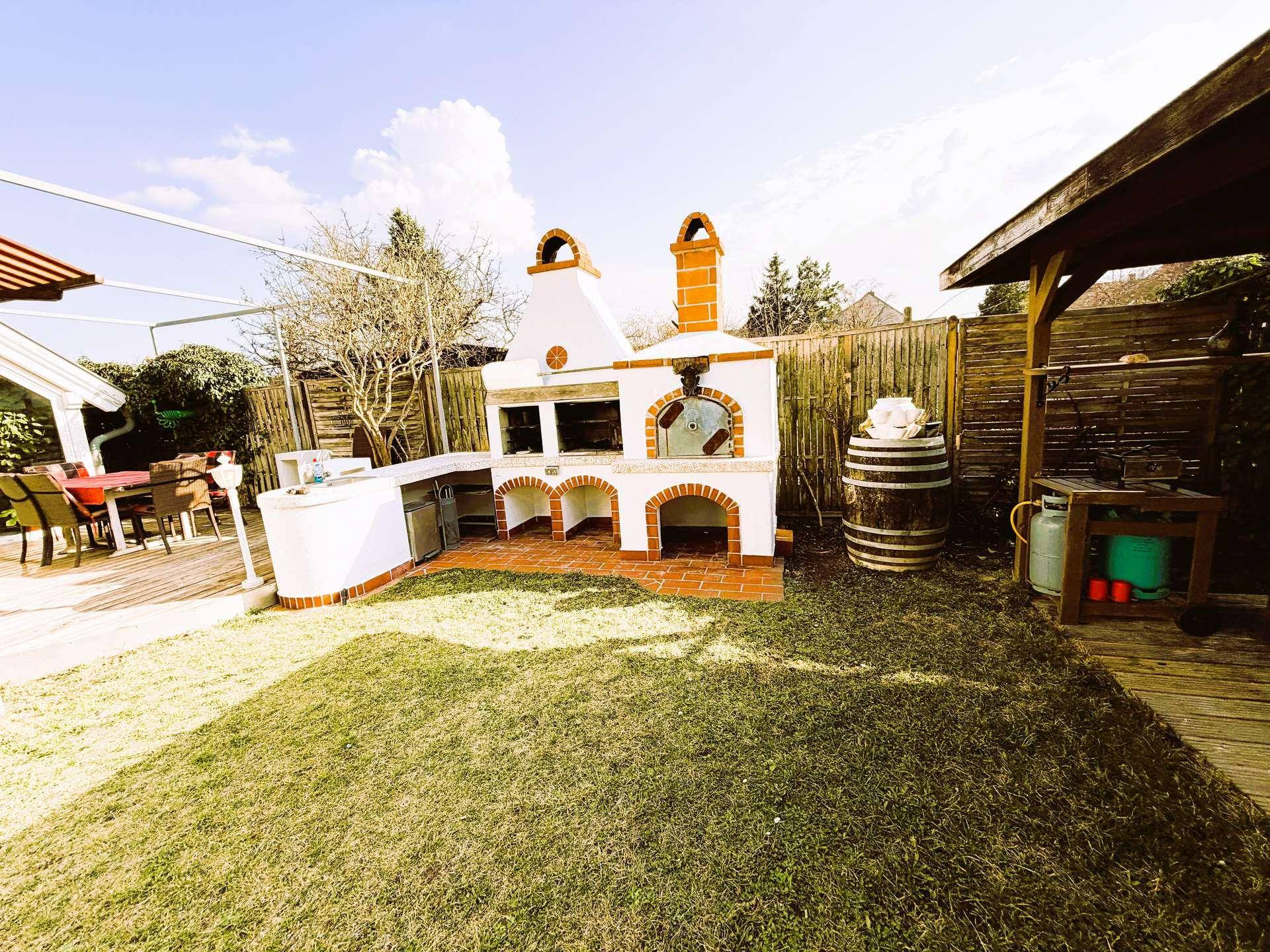 Gemauerte Außenküche mit Grill und Pizzaofen im grünen Garten, neben einem Holzpavillon.