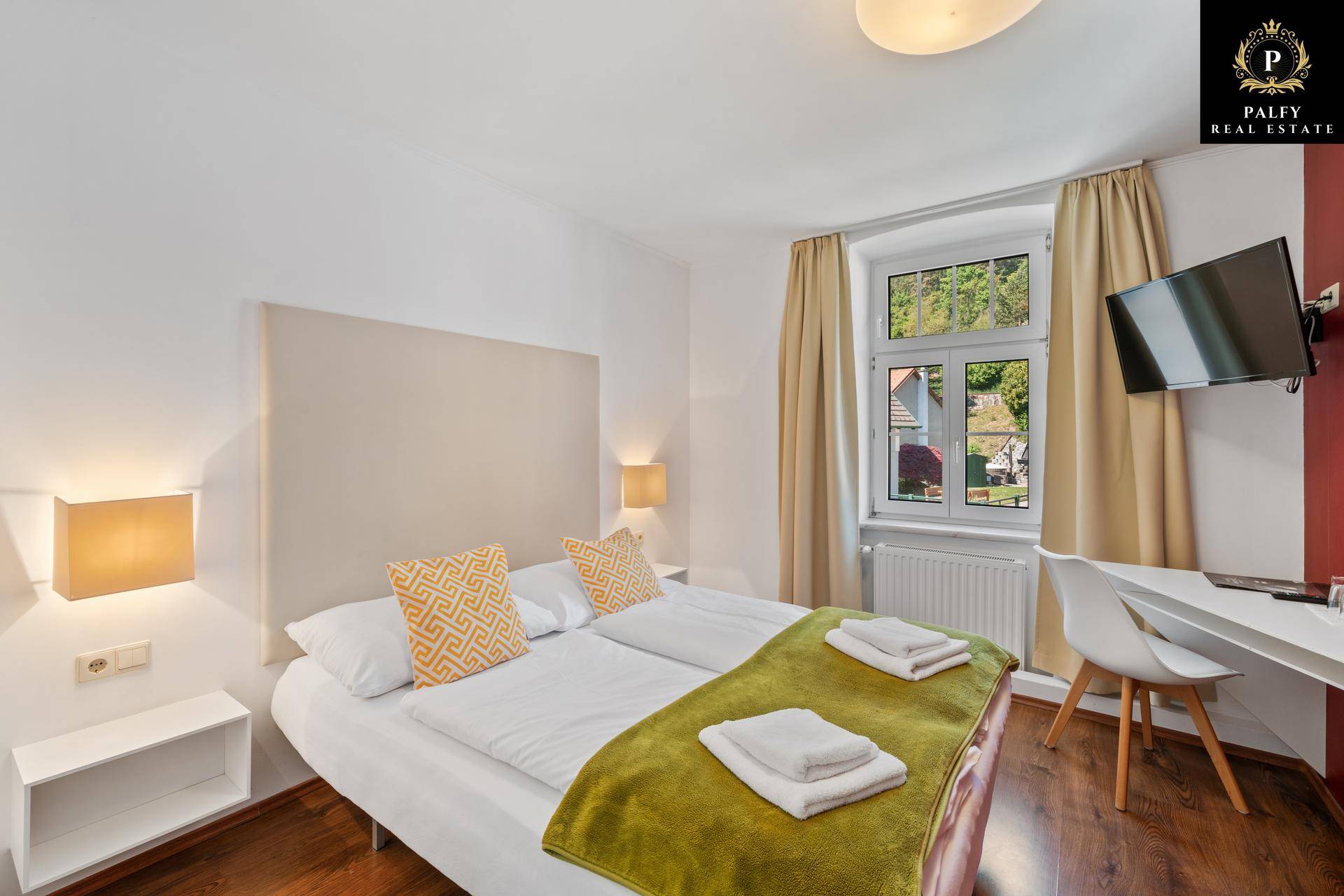 Helles Schlafzimmer mit Doppelbett, Schreibtisch und Fernseher, Holzboden und Fenster.