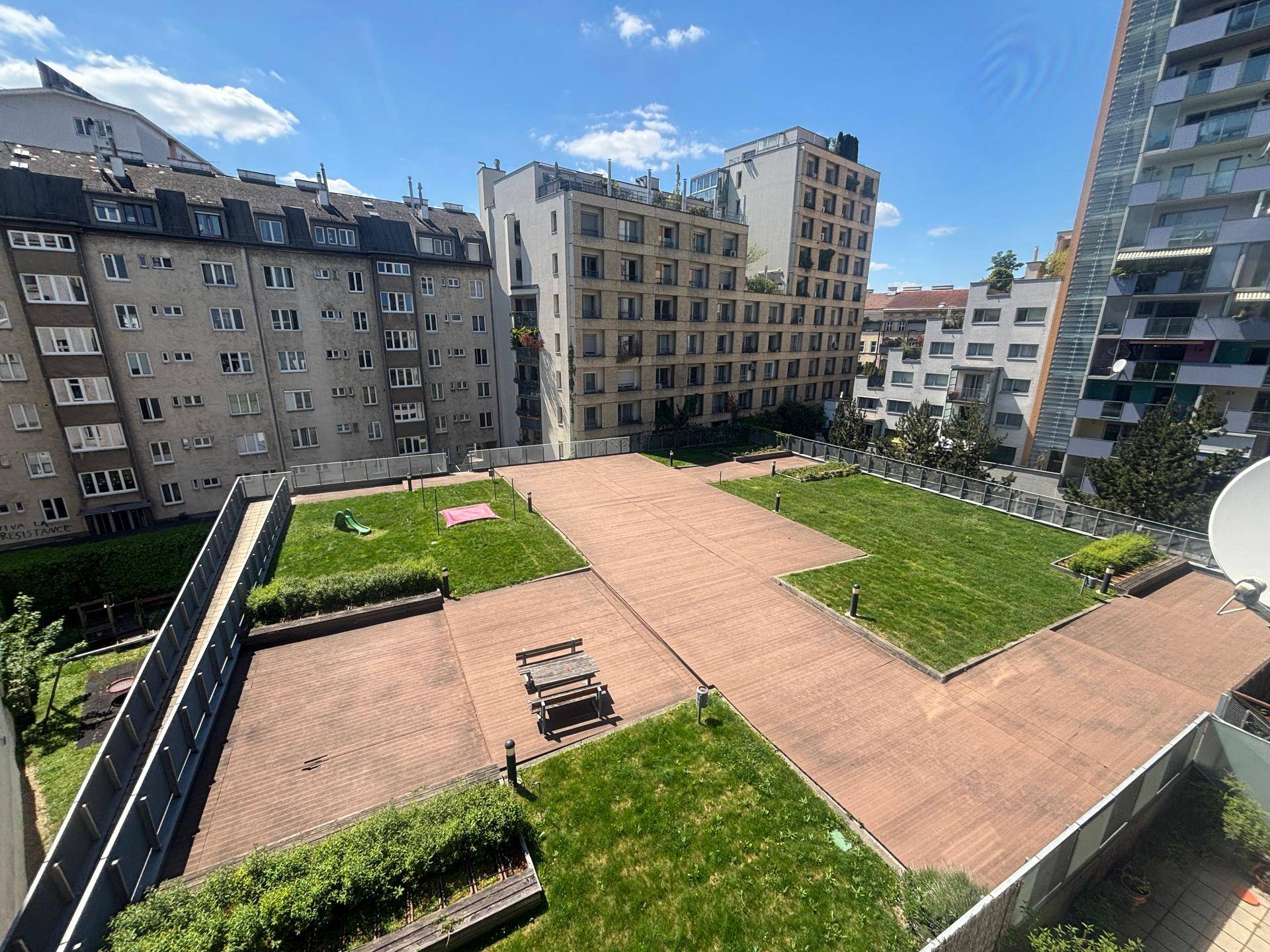 Weitläufige Dachterrasse mit Holzbelag, Grünflächen und Sitzgelegenheiten in urbaner Umgebung.