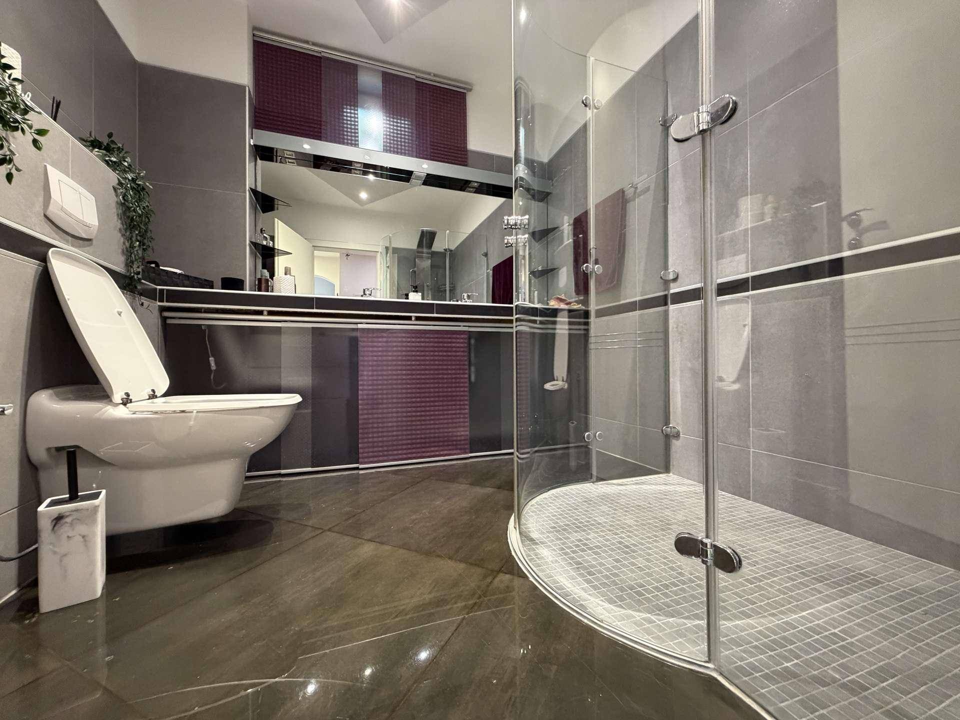 Modernes Badezimmer mit grauen Fliesen, wandhängendem WC und einer geräumigen Glasdusche.