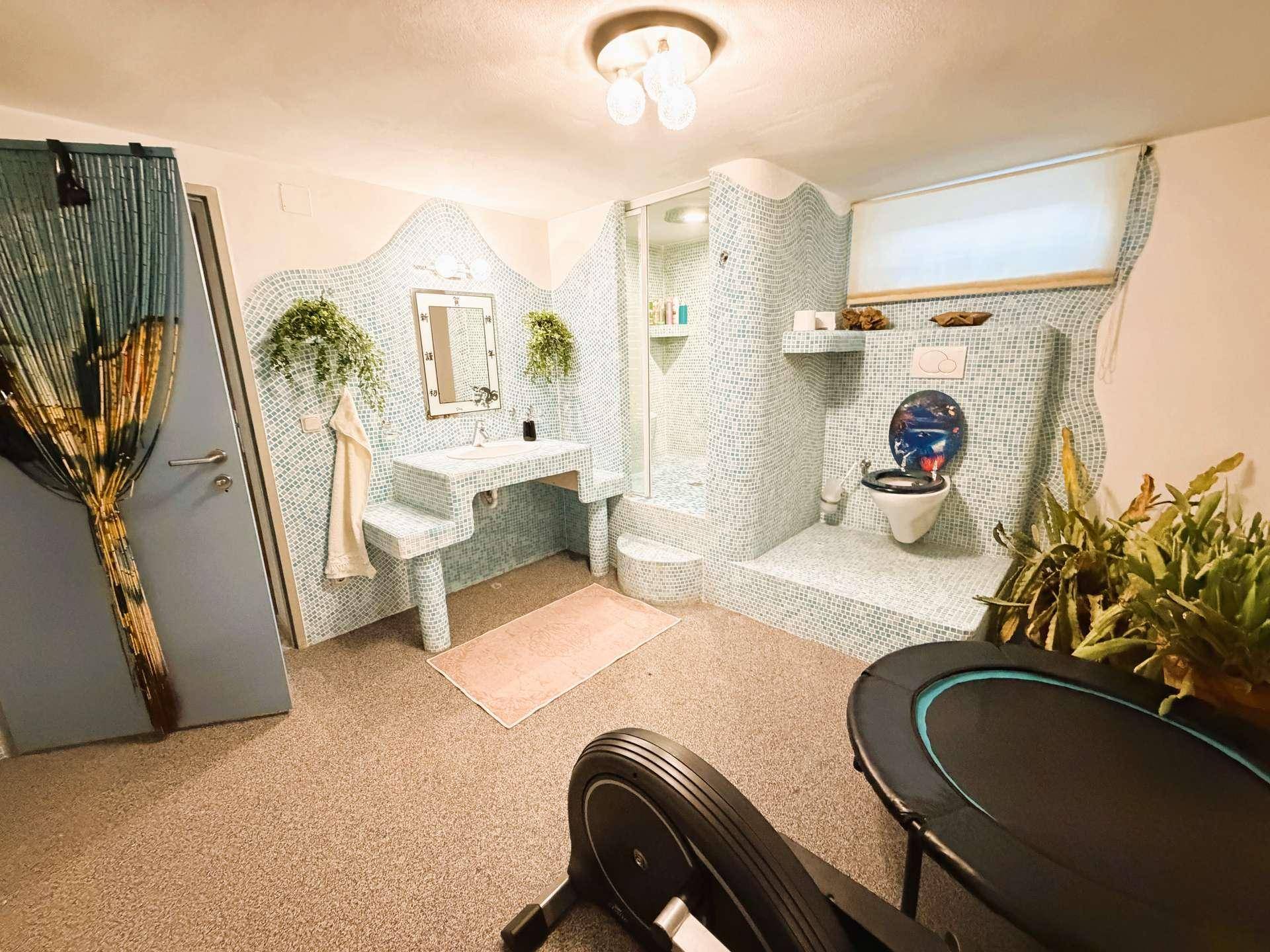 Kreativ gestaltetes Badezimmer im Keller mit Mosaikfliesen, Dusche, Toilette und Fitnessgeräten.