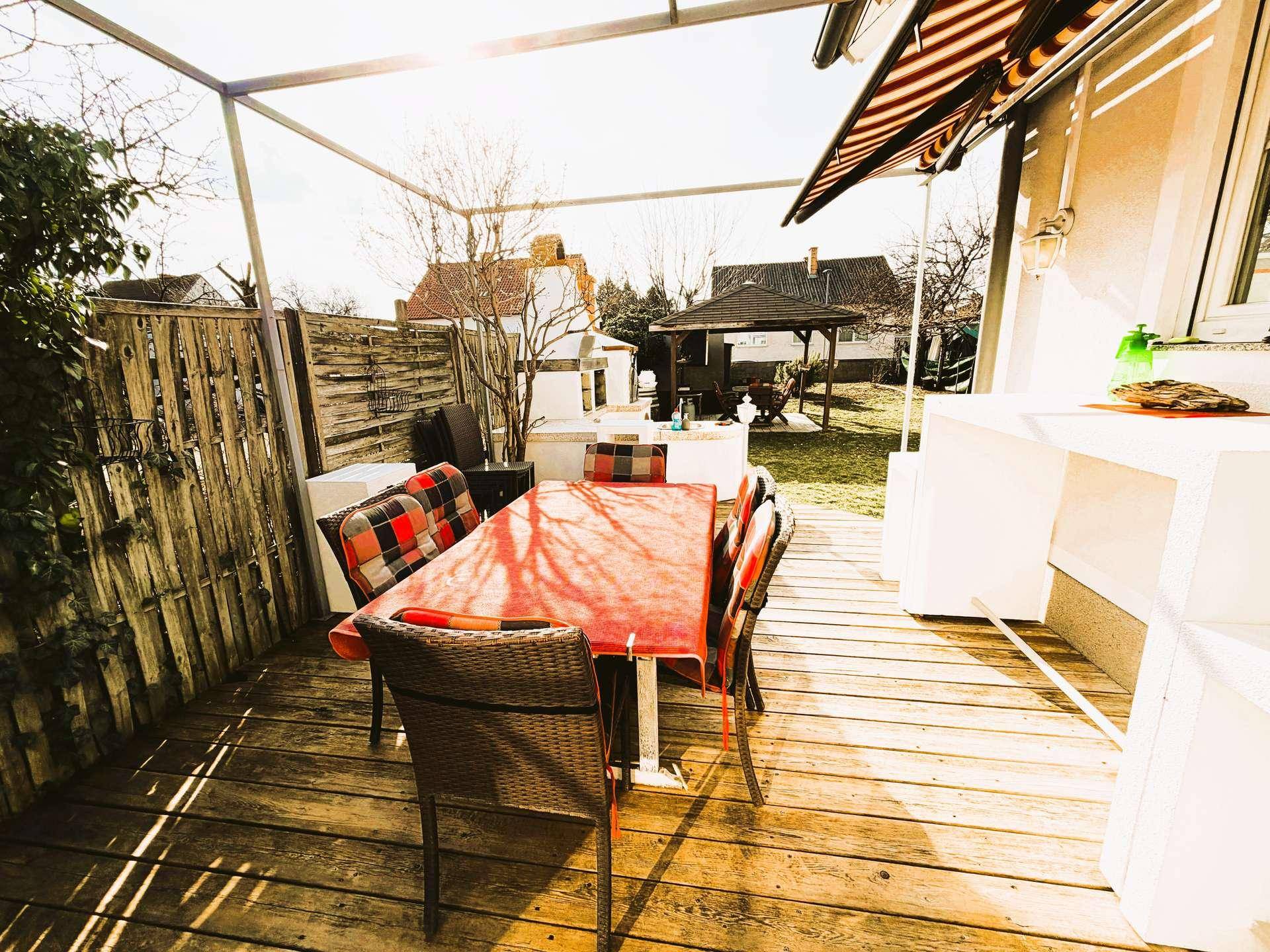 Sonniger Terrassenbereich mit Holzboden, Esstisch und Blick auf den Garten mit Pavillon.