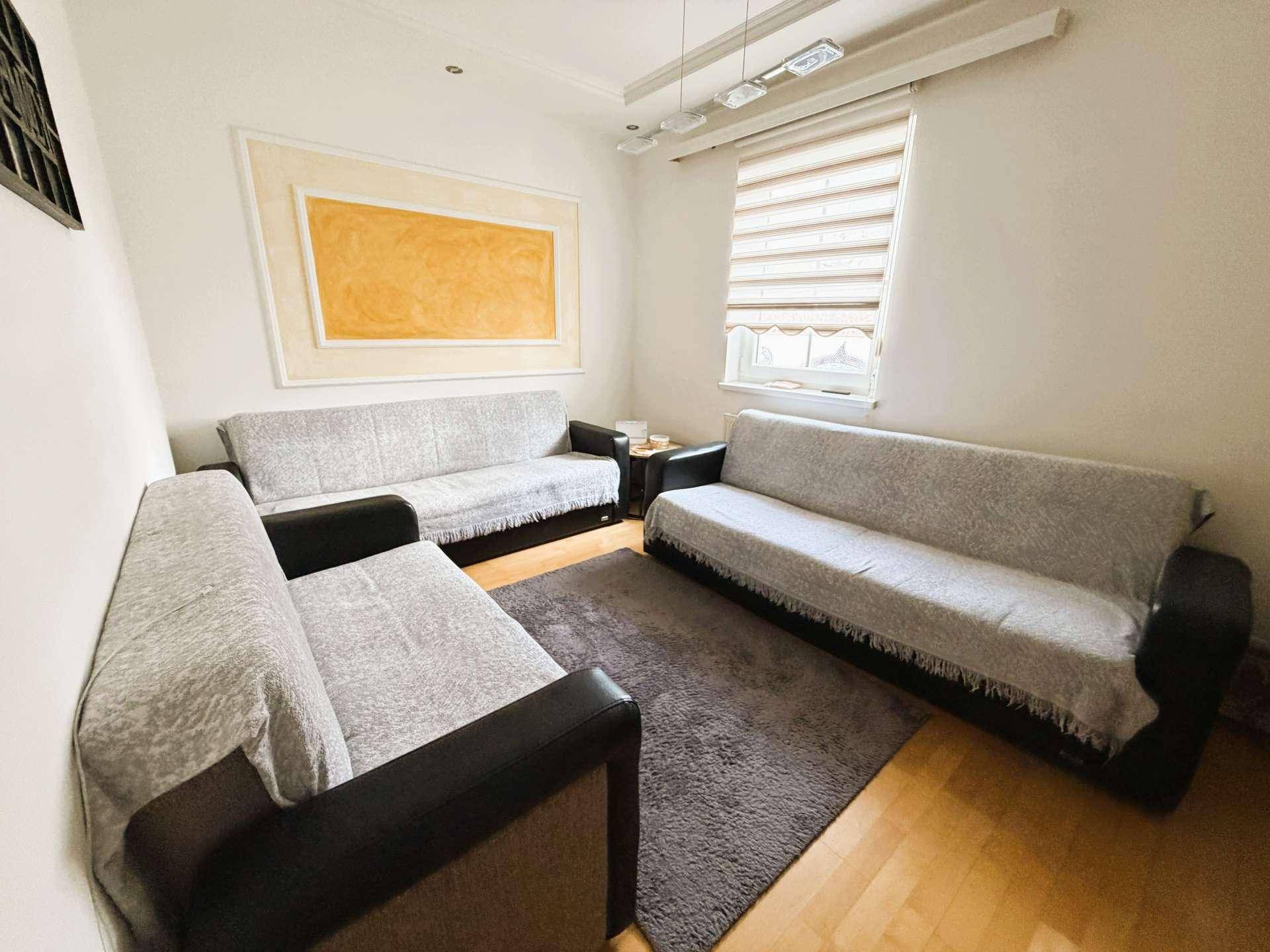 Gemütlicher Wohnraum mit drei Sofas, Teppichboden und einem Fenster mit Jalousien.