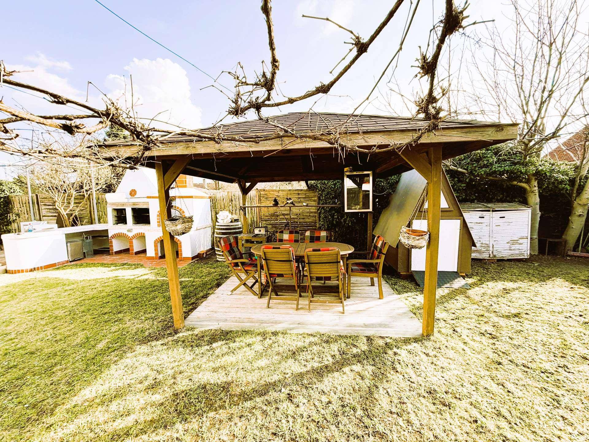 Großer Garten mit Holzpavillon, Esstisch und einer gemauerten Außenküche mit Grill.