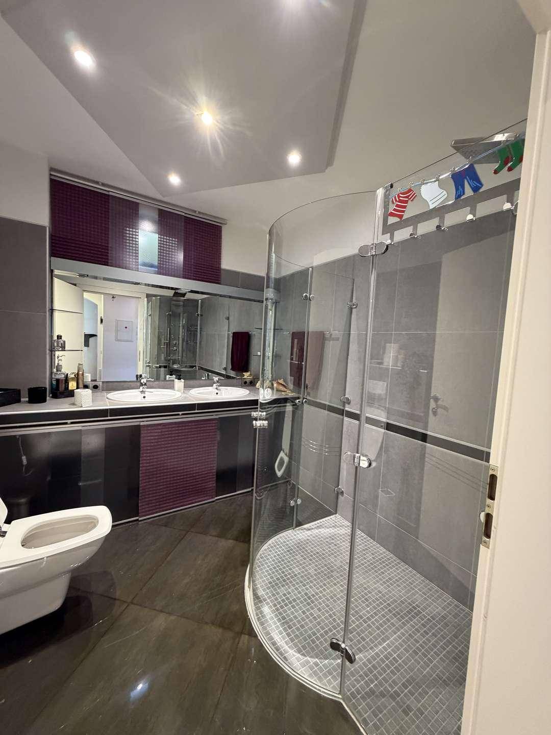 Modernes Badezimmer mit Doppelwaschbecken, Toilette und einer eleganten Glasduschkabine in Grau- und Lilatönen.