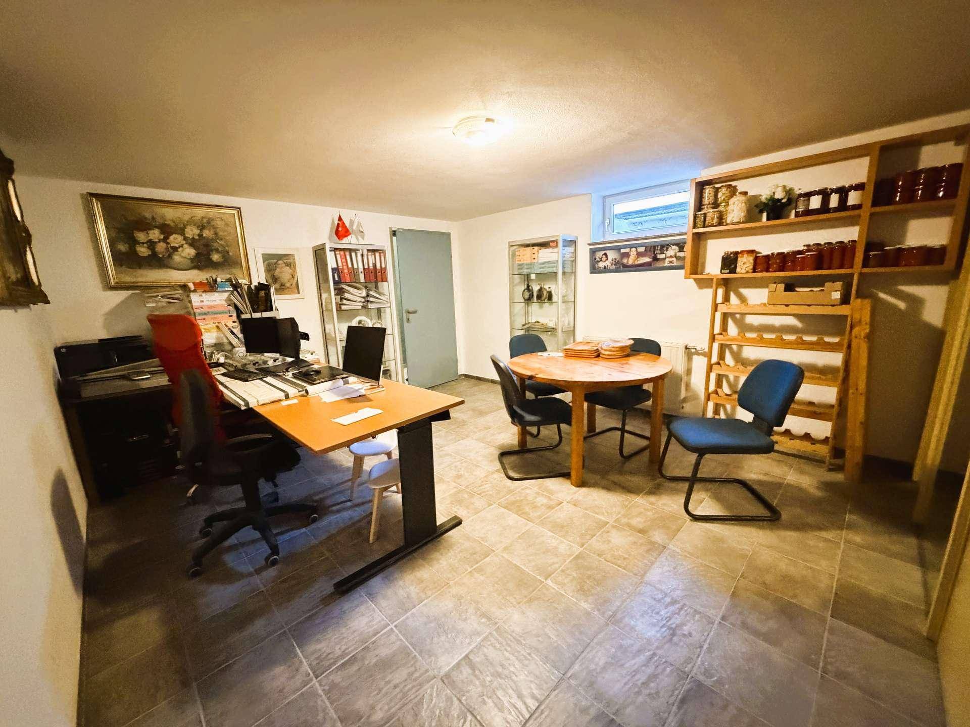 Funktionaler Raum im Kellergeschoss, eingerichtet als Büro mit Schreibtisch, Regalen und kleinem Esstisch.