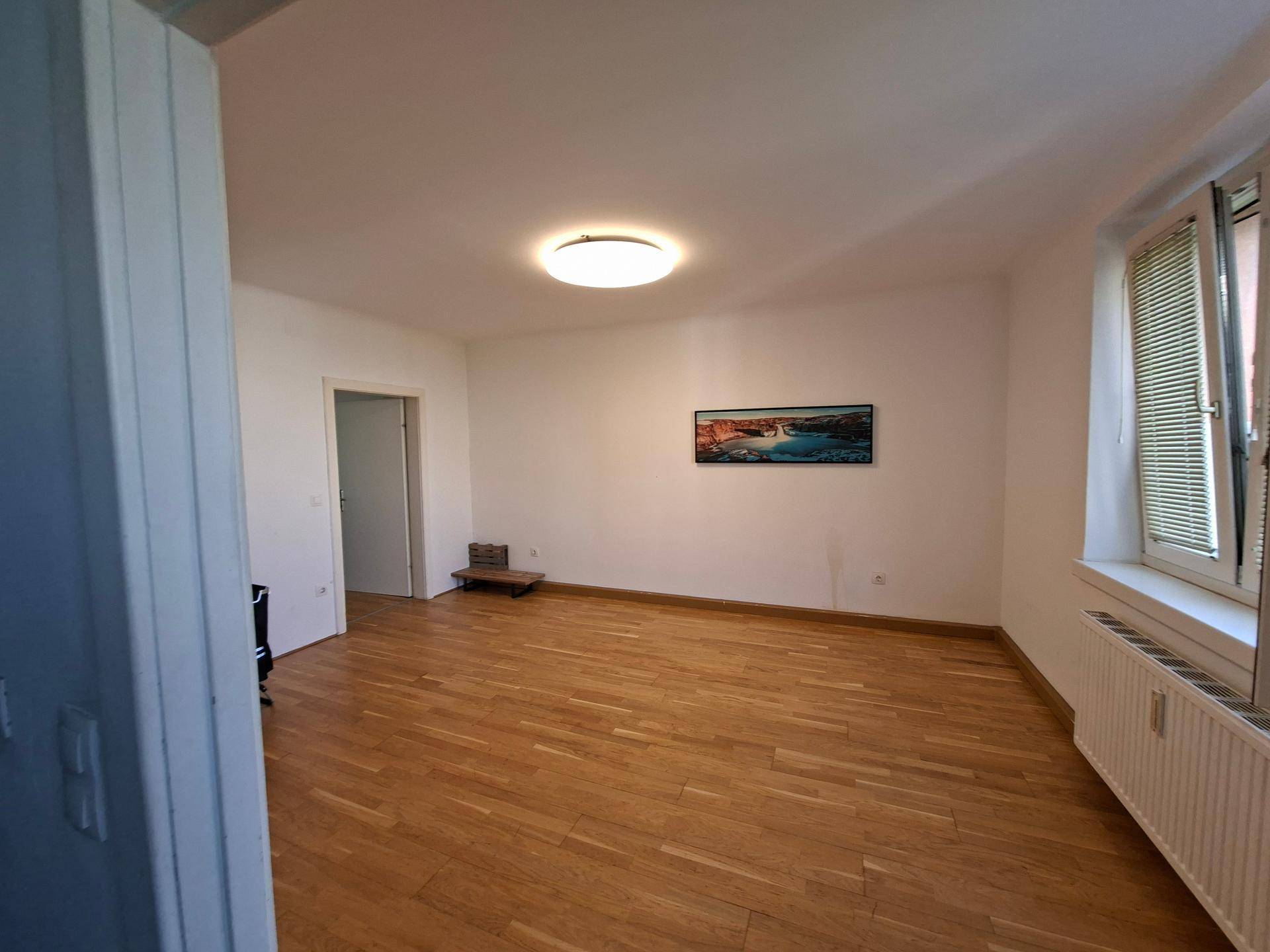 Leeres Wohnzimmer mit Holzboden, weißen Wänden und einem Fenster mit Heizkörper.
