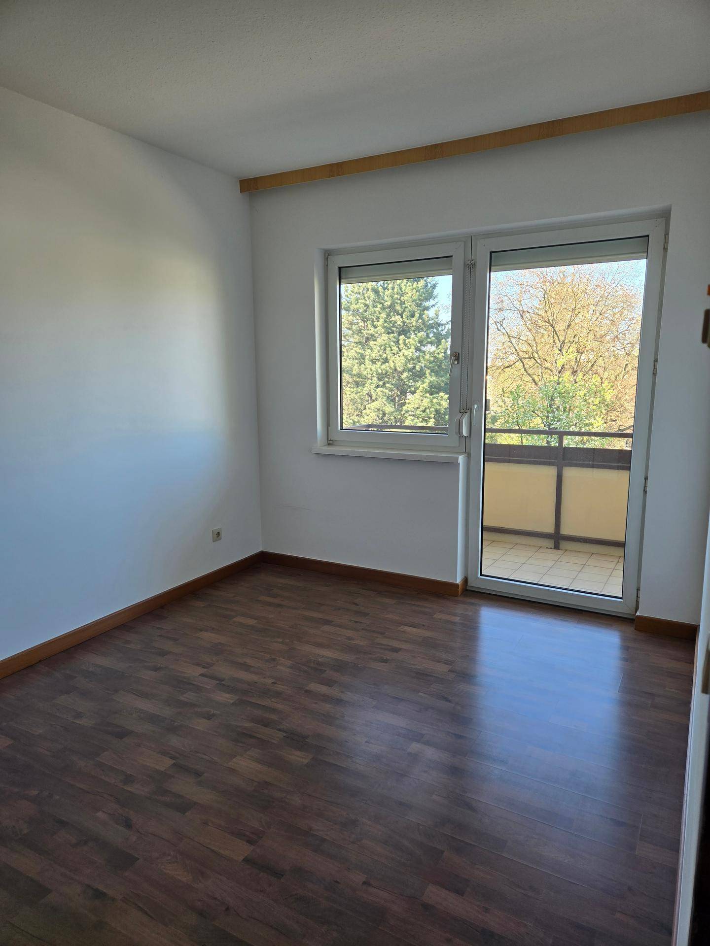 Schlafzimmer mit dunklem Laminatboden und Zugang zu einem Balkon mit Grünblick.