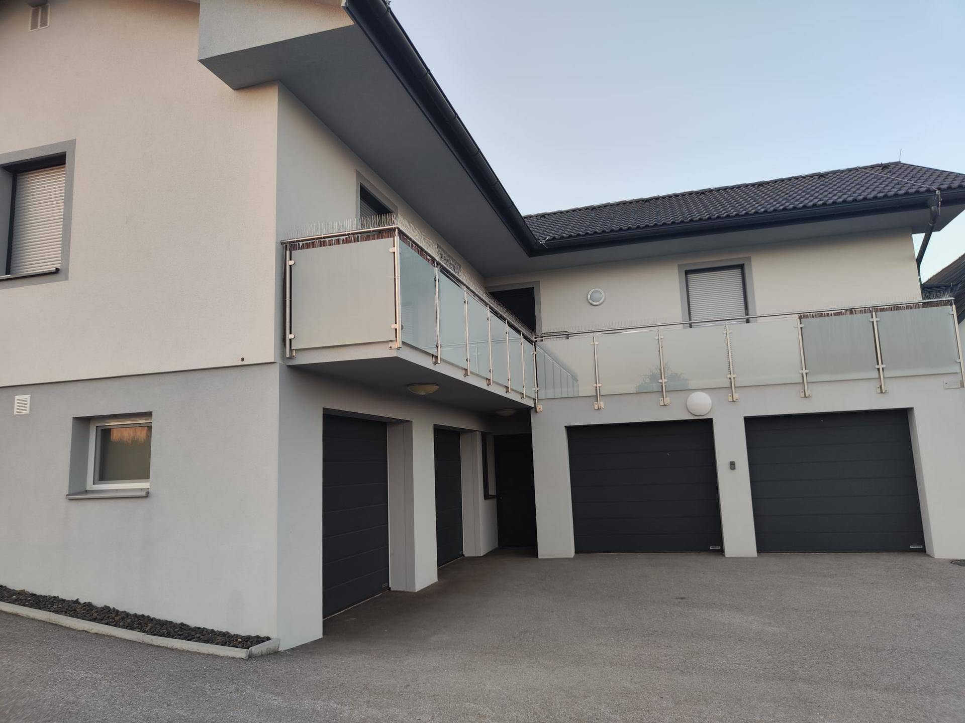 Moderne Hausfassade mit drei Garagen, Balkon mit Glasgeländer und Rollläden an den Fenstern.