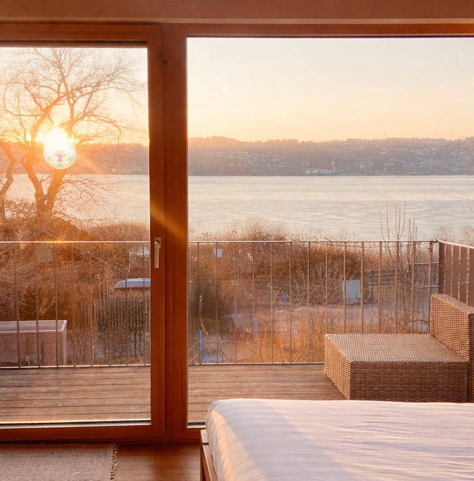 Helles Schlafzimmer mit Seeblick und direktem Zugang zu einer Holzterrasse bei Sonnenuntergang.