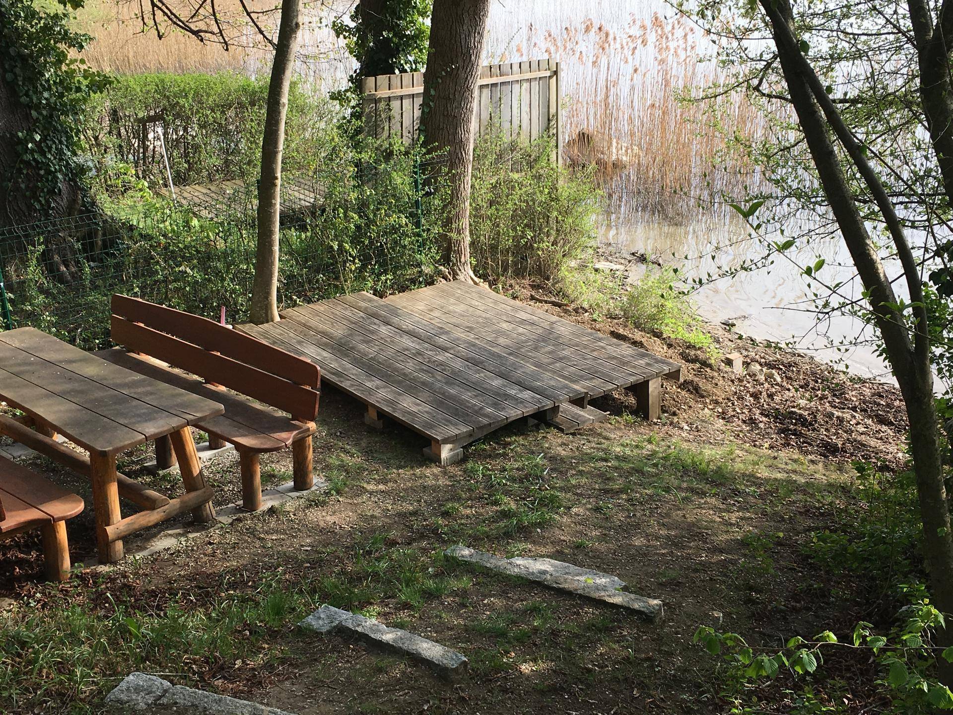 Rustikaler Sitzbereich am Seeufer mit Holztisch und Bänken unter Bäumen.