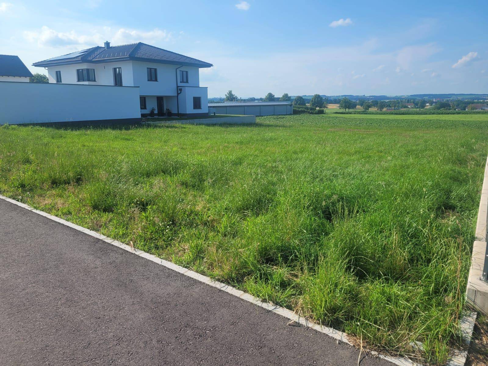 Unbebautes Grundstück mit hohem Gras und Neubau in ländlicher Umgebung.