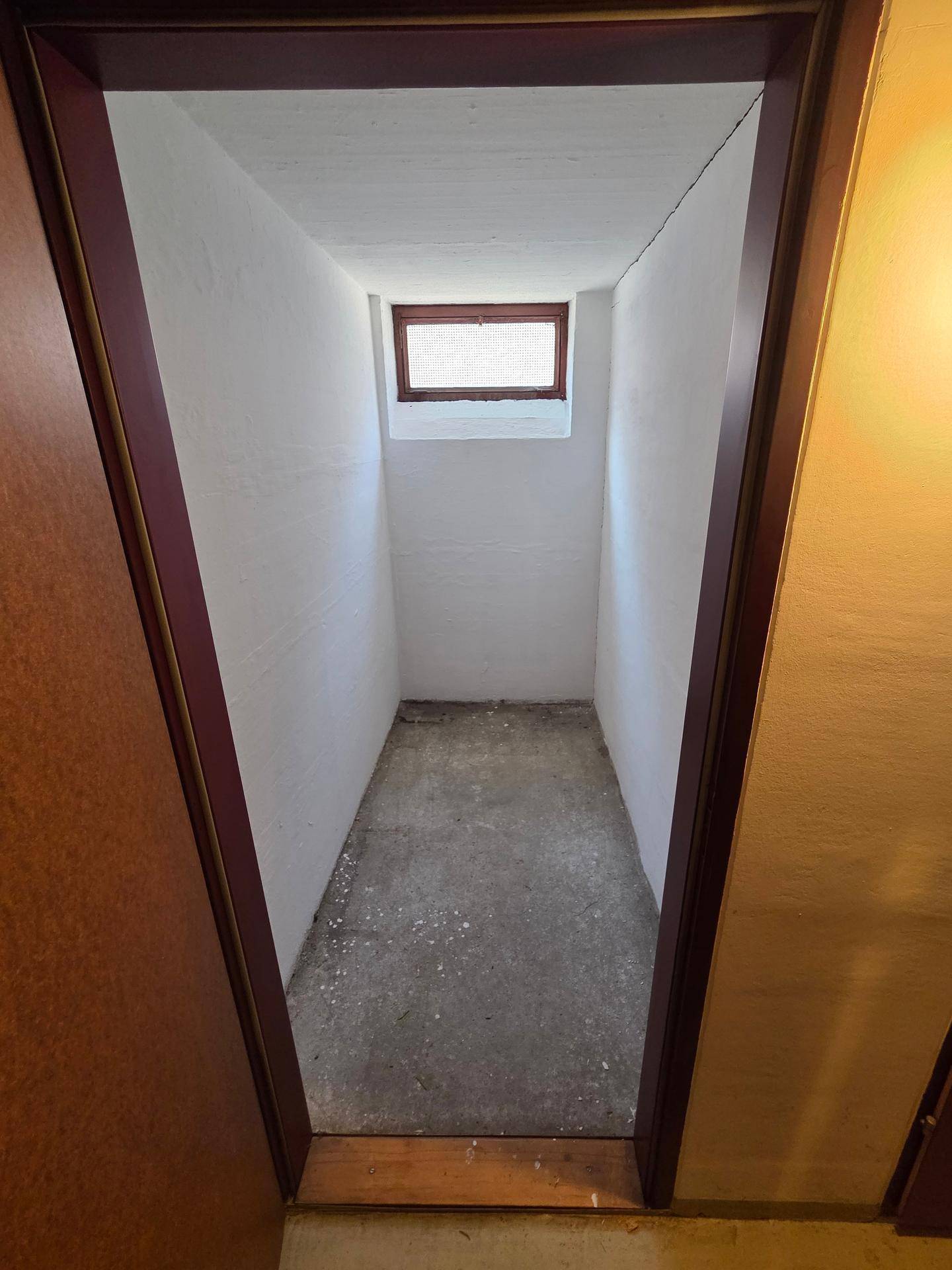 Schmaler Abstellraum oder Kellerabteil mit kleinem Fenster und Betonboden.