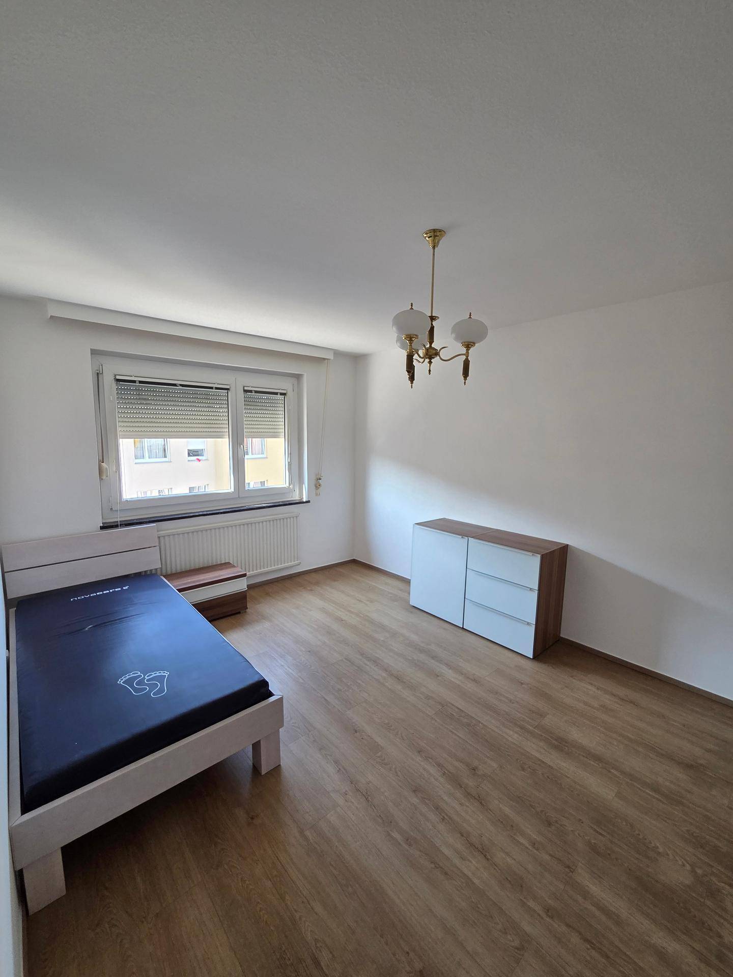 Schlafzimmer mit Holzboden, Einzelbett, Kommode und Fenster mit Rollladen.