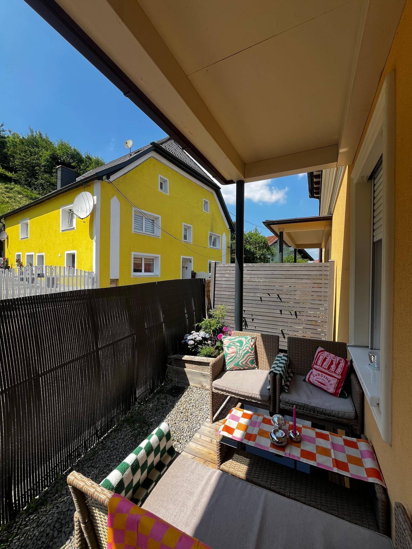 Gemütliche Terrasse mit Korbmöbeln, bunten Kissen und Blick auf ein gelbes Haus in der Ferne.