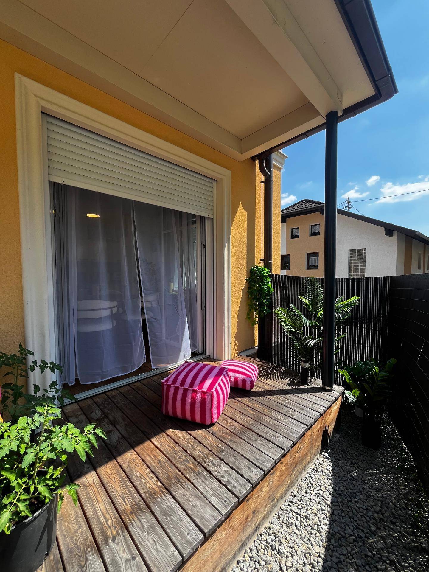 Sonniger Terrassenbereich mit Holzdeck, Pflanzen und pinken Sitzkissen für entspannte Stunden im Freien.