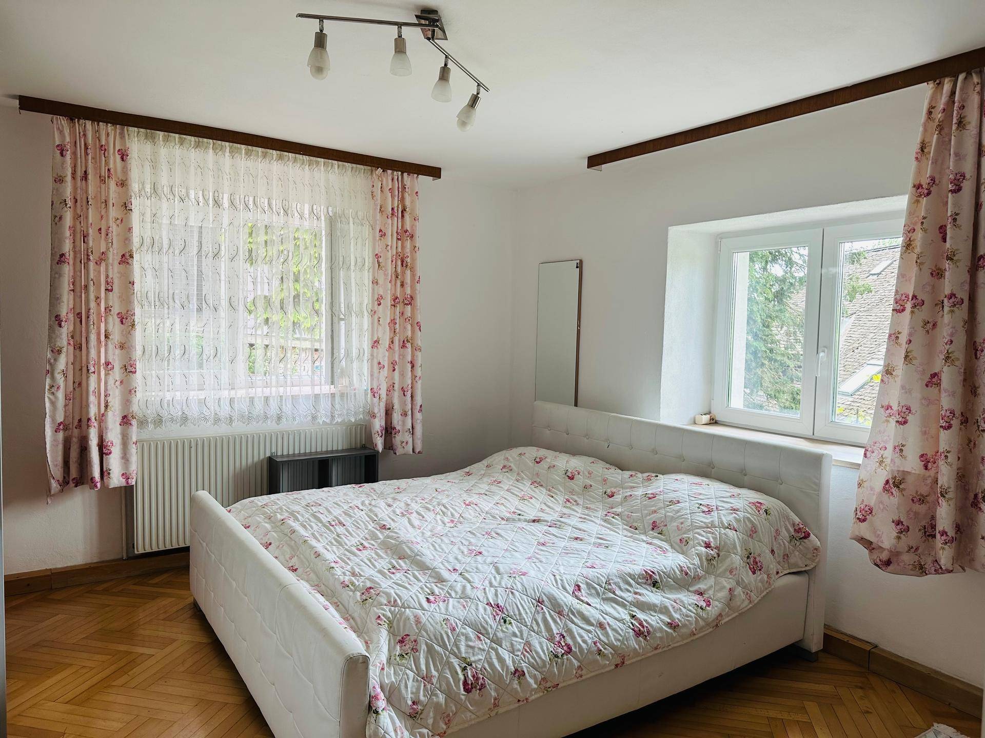 Helles Schlafzimmer mit weißem Bett, floralen Vorhängen und klassischem Parkettboden.