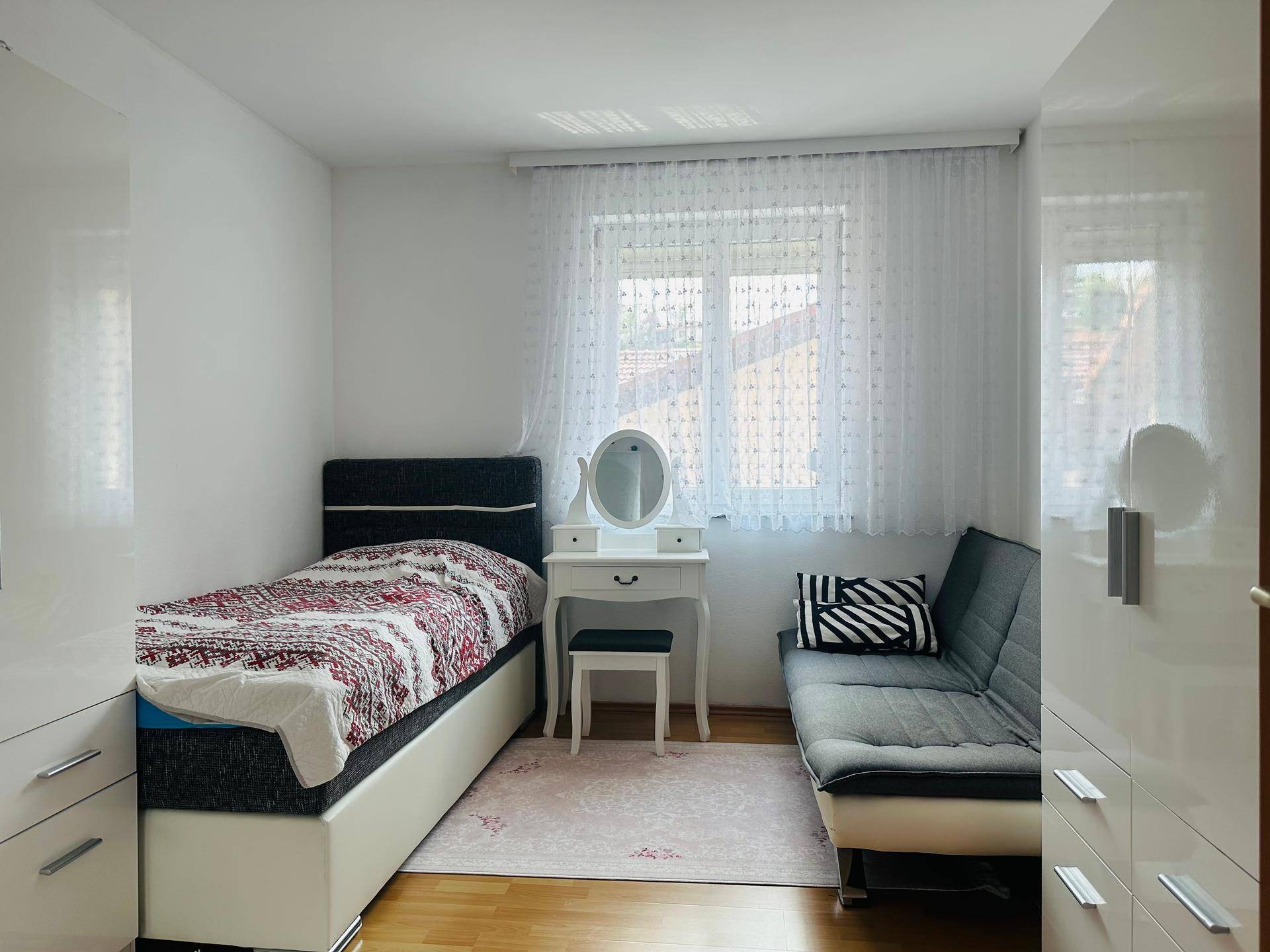 Helles Schlafzimmer mit Einzelbett, Schminktisch und kleiner Couch, modern eingerichtet.