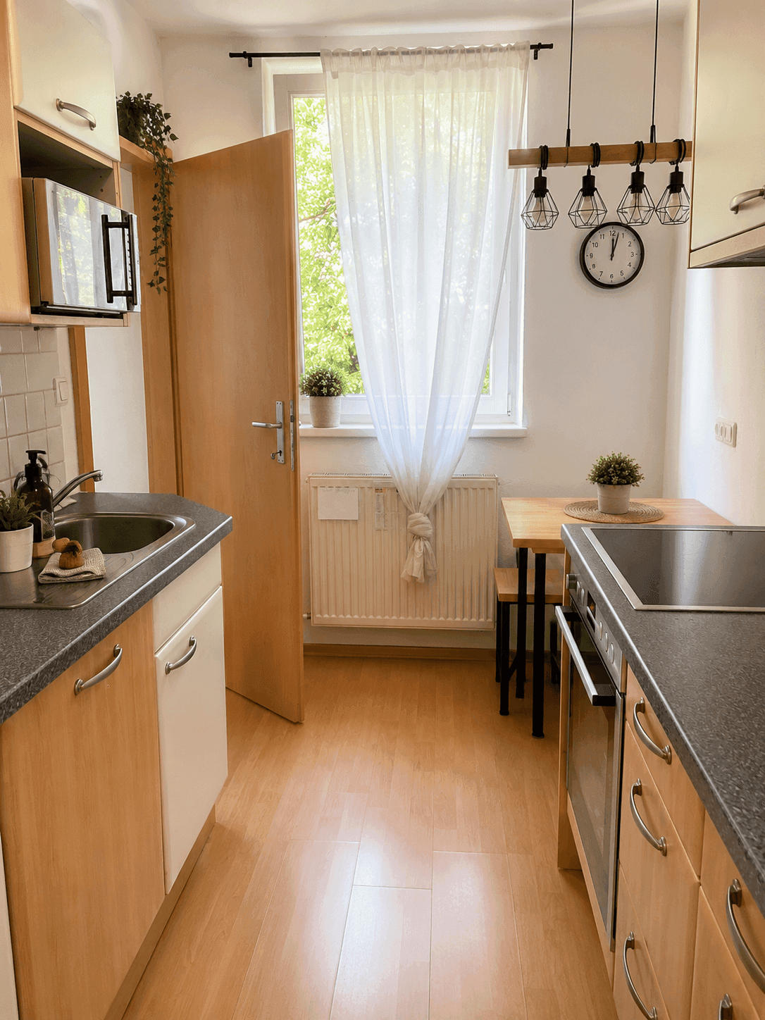 Funktionale Küche mit hellen Holzfronten, kleinem Esstisch und Fenster mit Vorhang.
