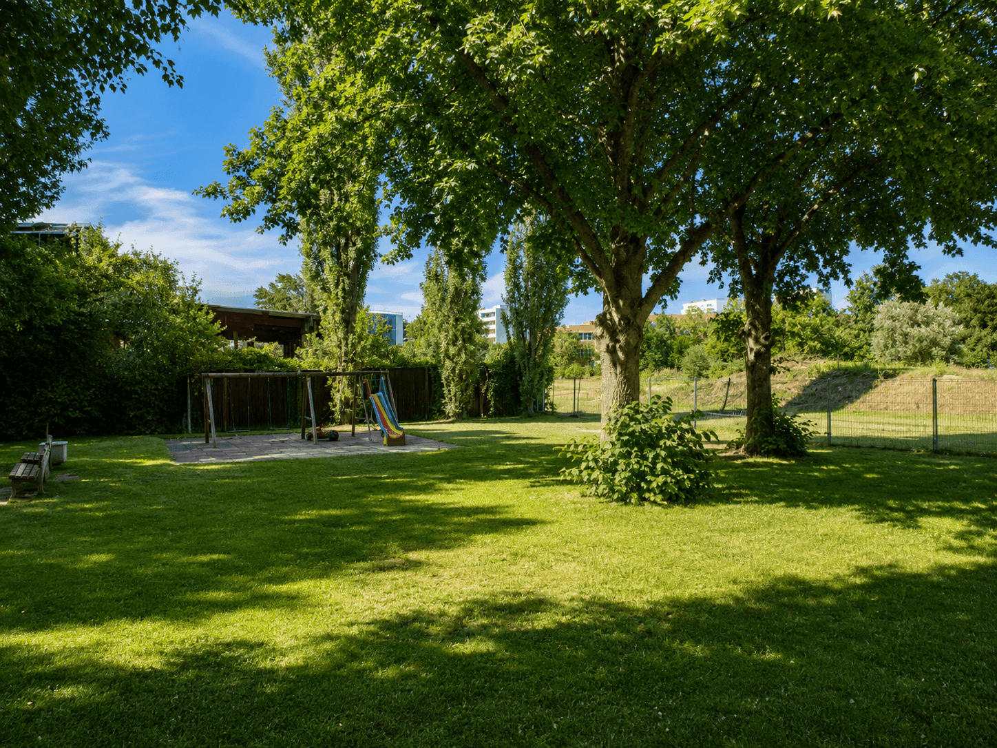 Großer Gemeinschaftsgarten mit Spielplatz, Rasenfläche und hohen Bäumen unter blauem Himmel.