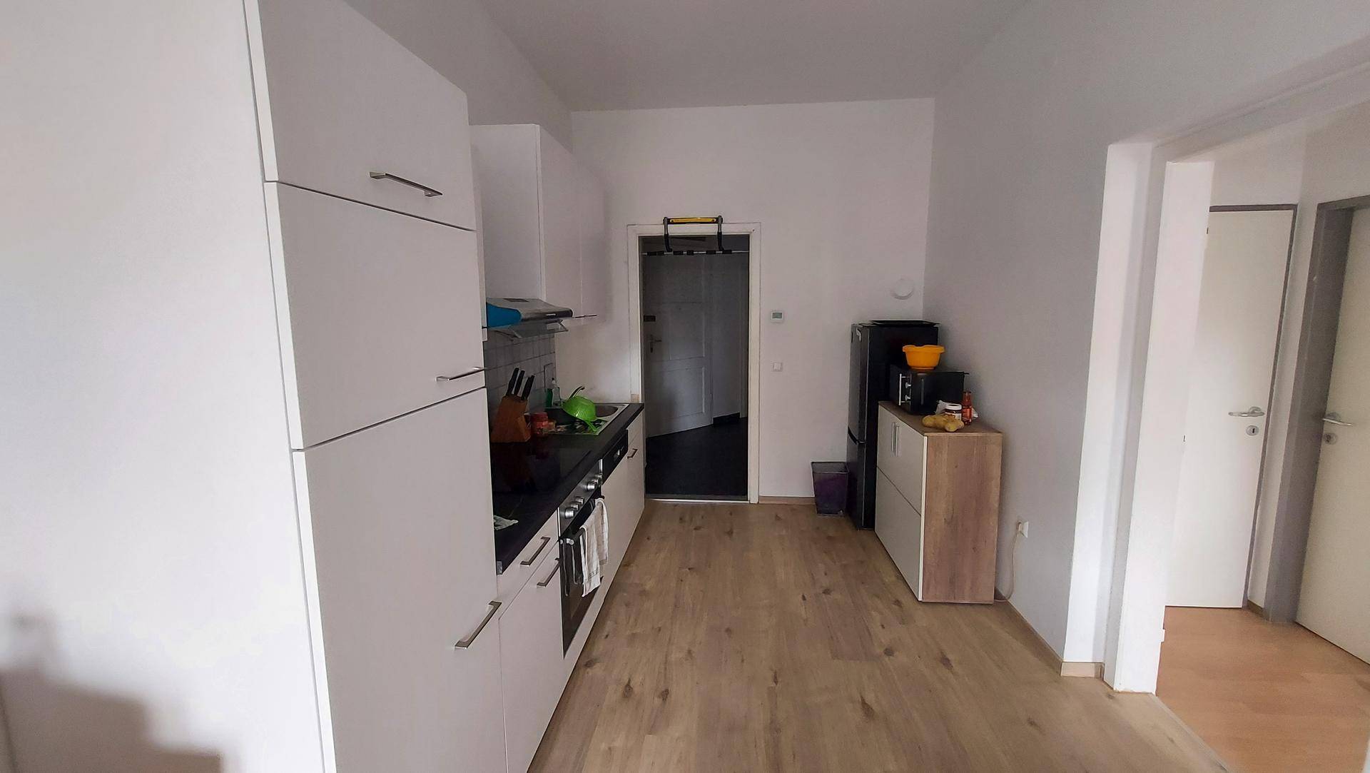 Längliche Küche mit weißen Schränken, Holzfußboden und Zugang zu weiteren Räumen.