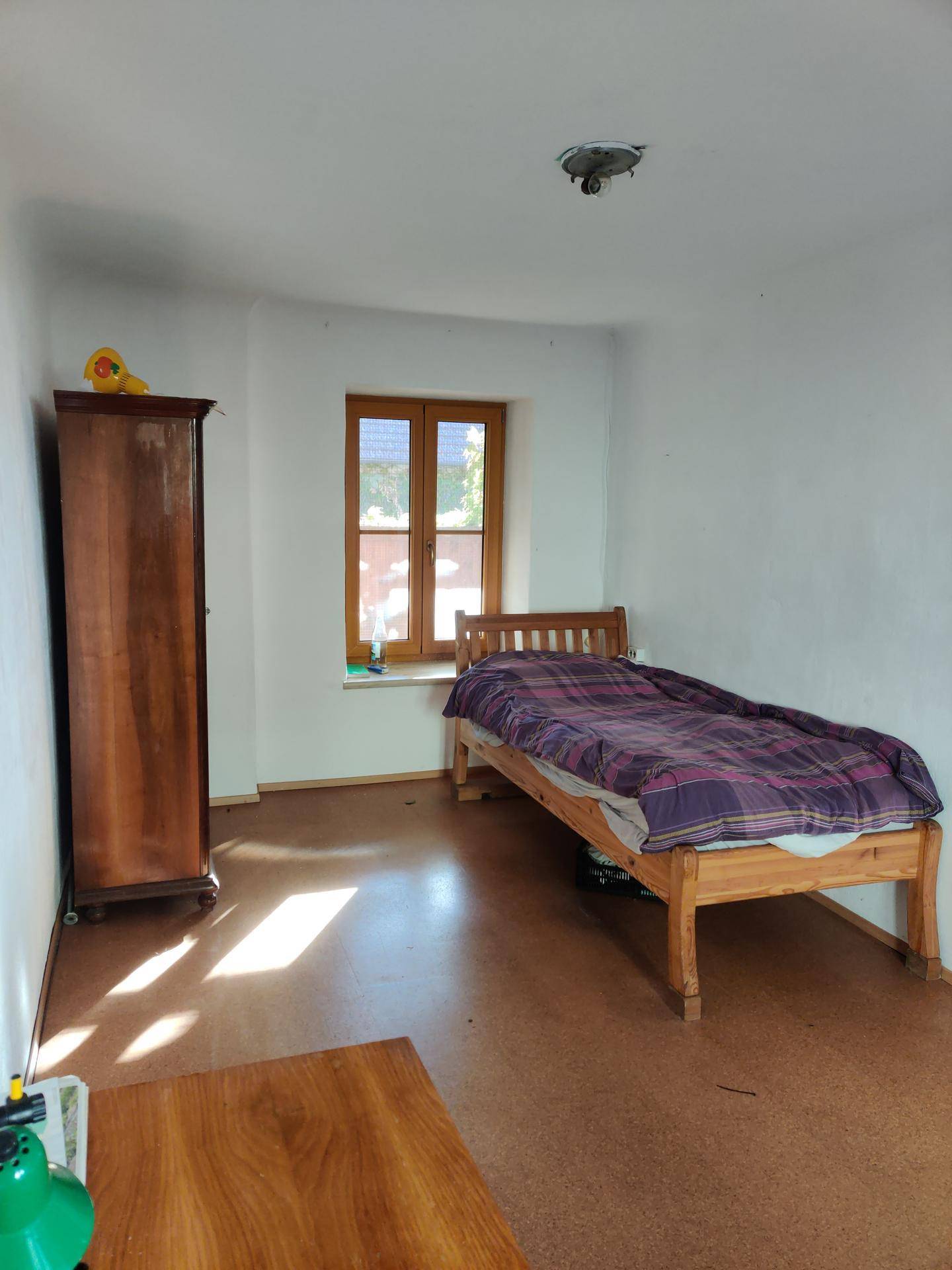 Schlafzimmer mit Holzbett, Kleiderschrank und einem Fenster, das Sonnenlicht hereinlässt.