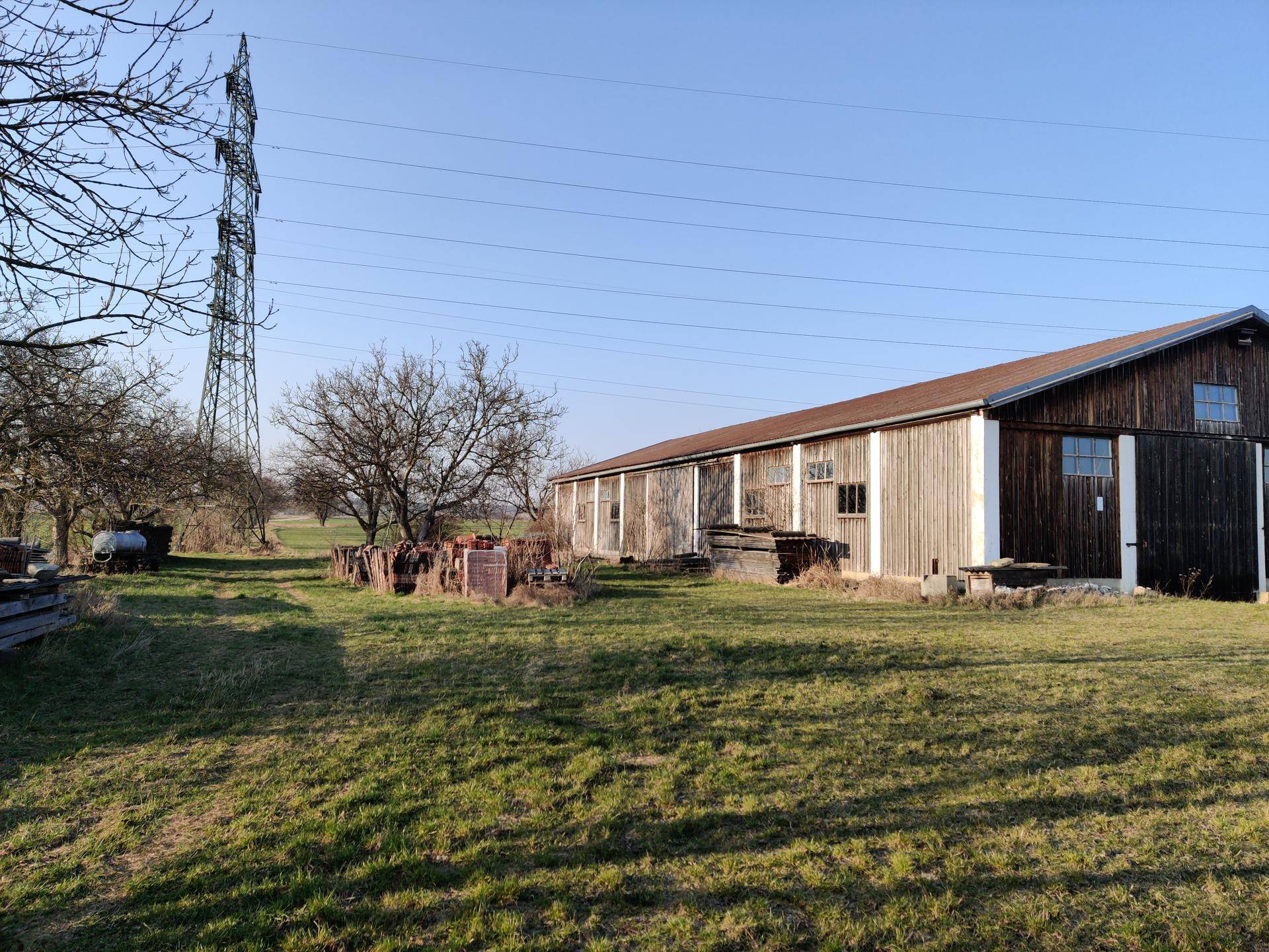 Langes Holzgebäude in ländlicher Umgebung mit weiten Feldern und einem Strommast im Hintergrund.