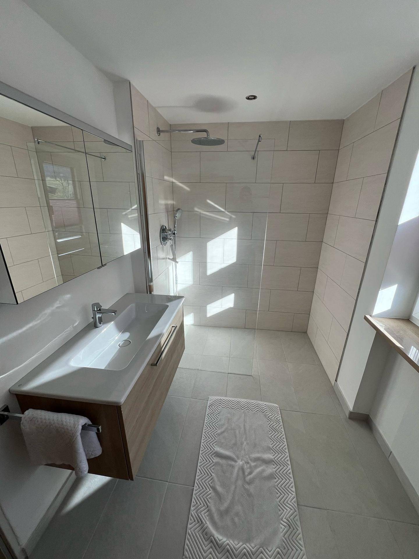 Modernes Badezimmer mit begehbarer Dusche, großem Waschbecken und Spiegelschrank, hell gefliest und mit Tageslicht.