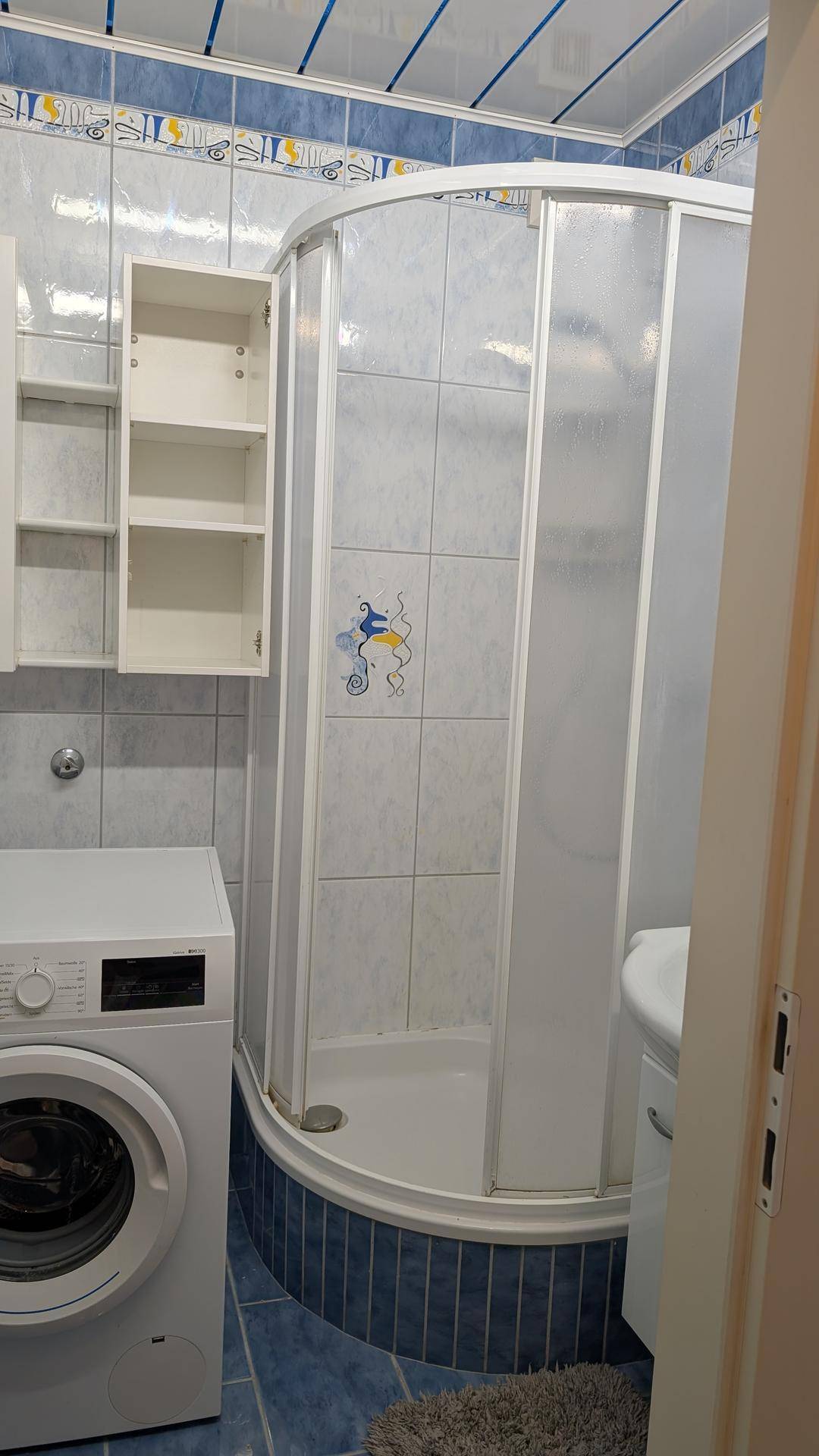 Badezimmer mit Duschkabine, Waschmaschine und blauen Bodenfliesen.