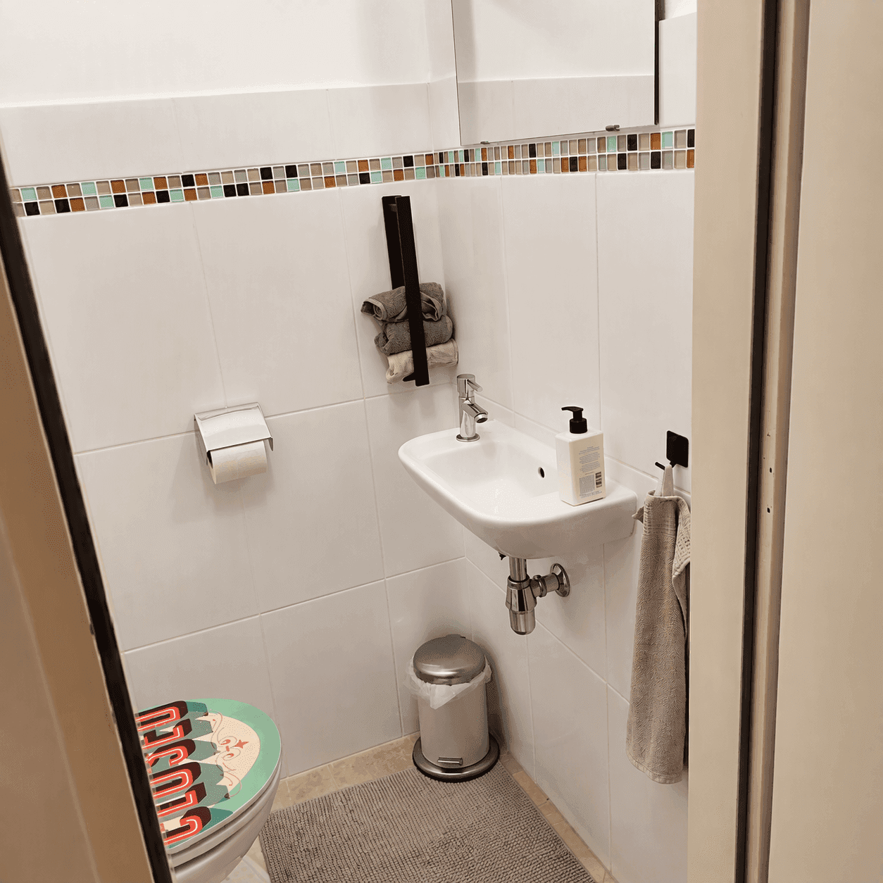 Kleines Gäste-WC mit weißen Fliesen, einem Waschbecken und einer farbigen Bordüre.