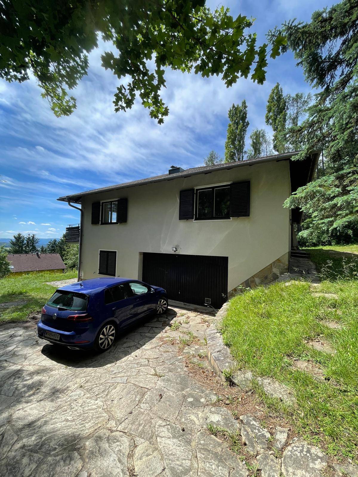 Haus mit Garage und Auffahrt, umgeben von viel Grün und Bäumen unter blauem Himmel.