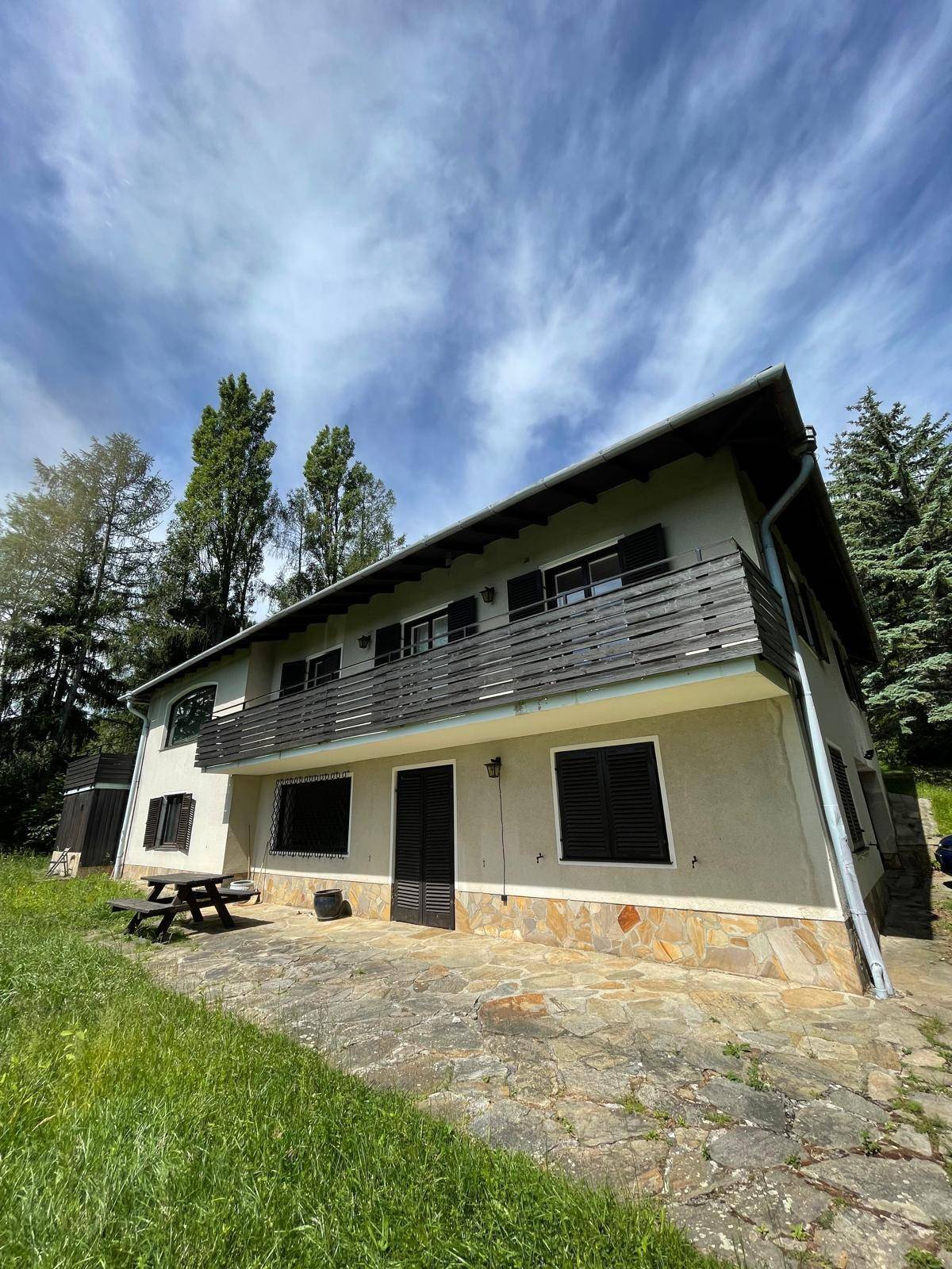 Zweistöckiges Haus mit Balkon und gepflasterter Terrasse, umgeben von Bäumen und Grünflächen.