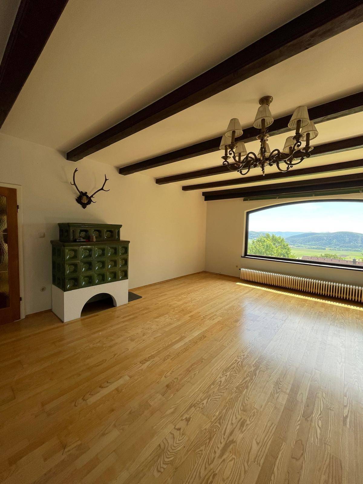 Helles Wohnzimmer mit Holzbalkendecke, Kachelofen und weitem Blick in die Landschaft.