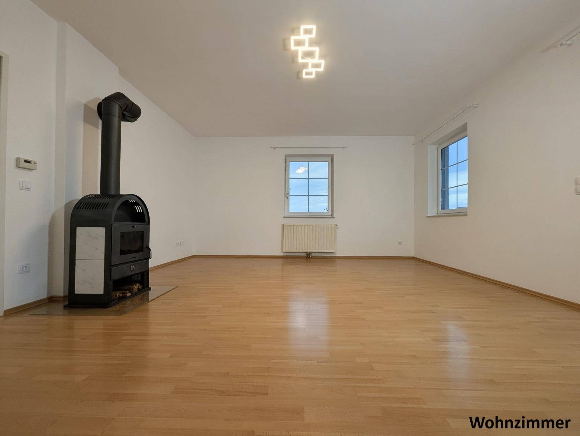 Helles Wohnzimmer mit Parkettboden, zwei Fenstern und einem modernen Holzofen für behagliche Wärme.