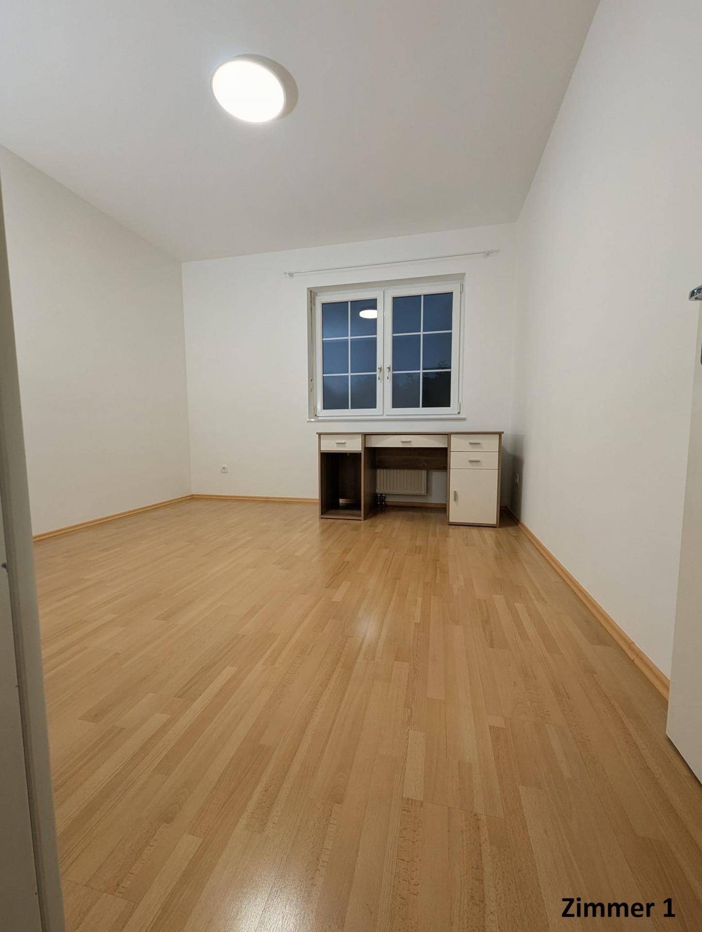Freundliches Zimmer mit Holzboden und großem Fenster, das viel Tageslicht hereinlässt.