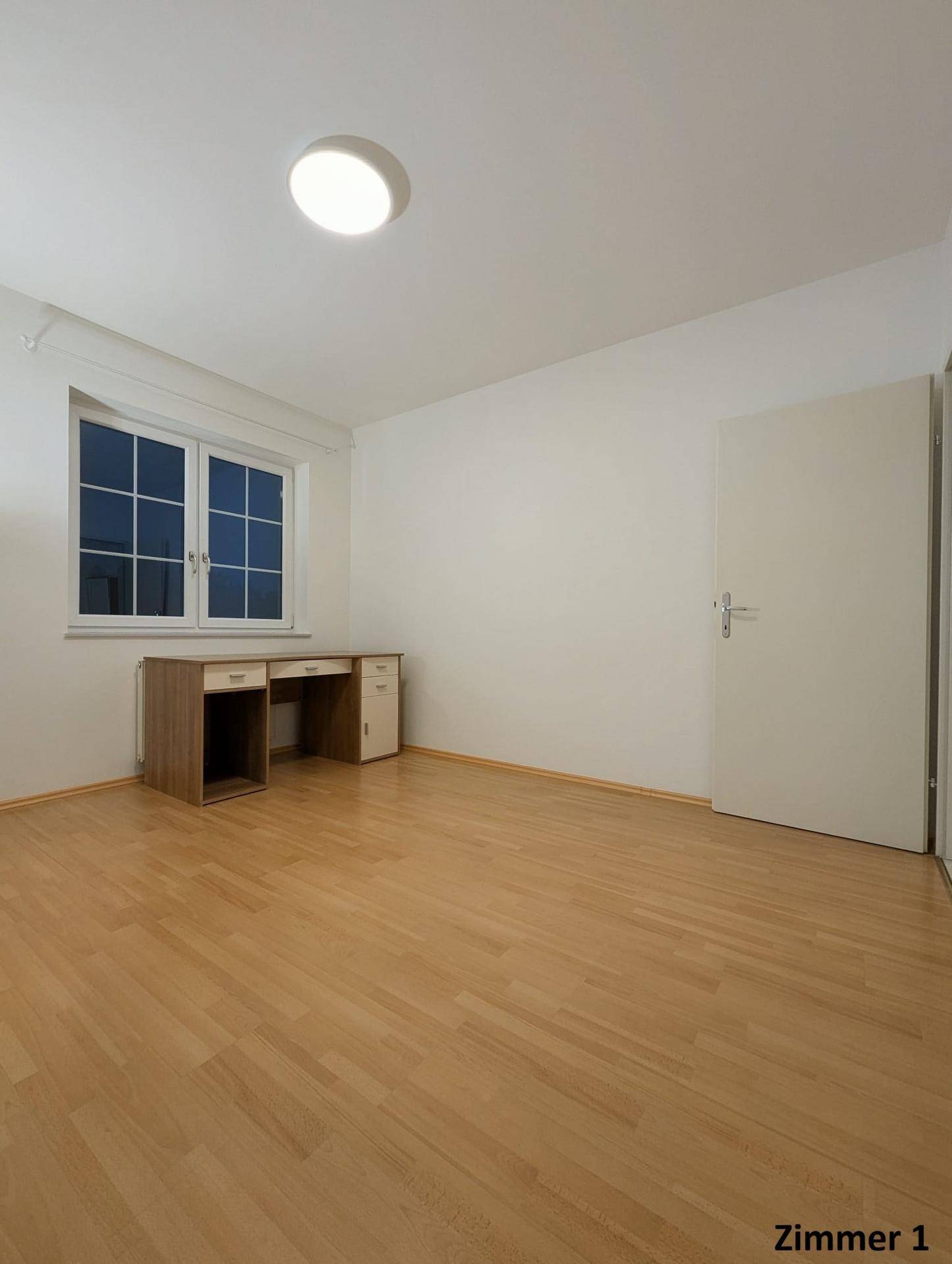 Helles Zimmer mit Holzboden, großem Fenster und einem Schreibtisch unter dem Fenster.