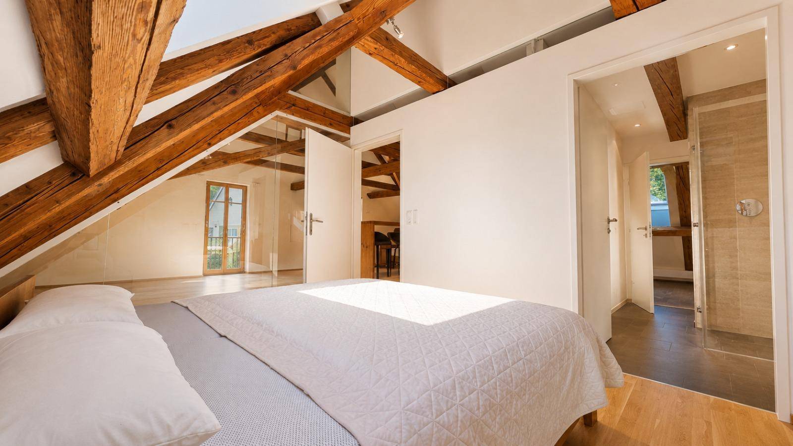 Helles Schlafzimmer im Dachgeschoss mit Holzbalken, gemütlichem Bett und Zugang zu einem weiteren Raum.