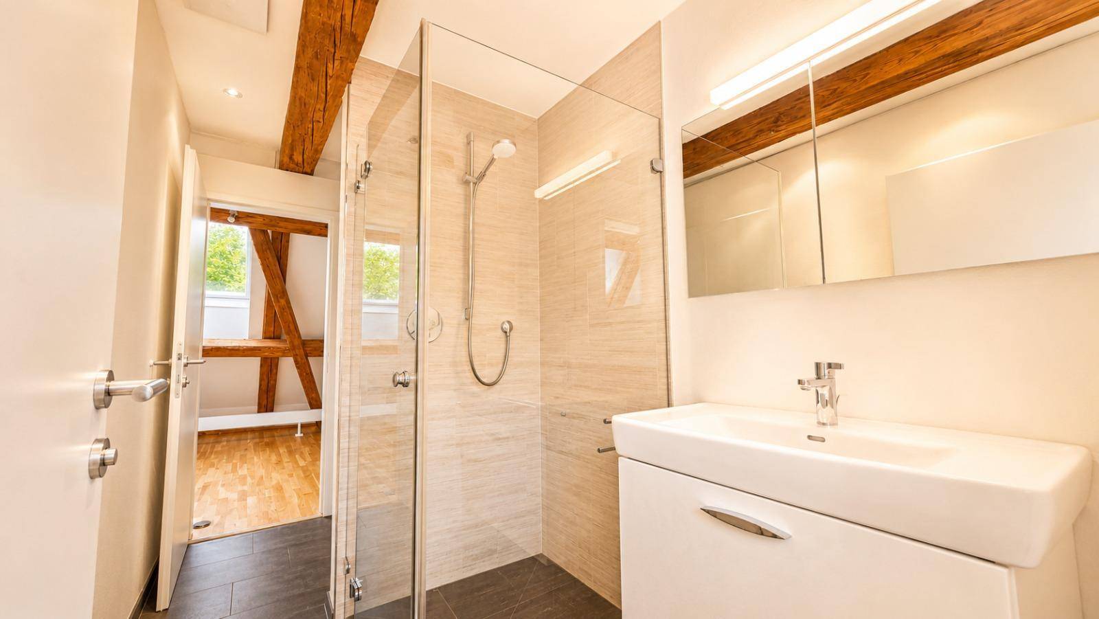 Modernes Badezimmer mit begehbarer Dusche, Einzelwaschbecken und dunklen Fliesen, durch Holzbalken akzentuiert.