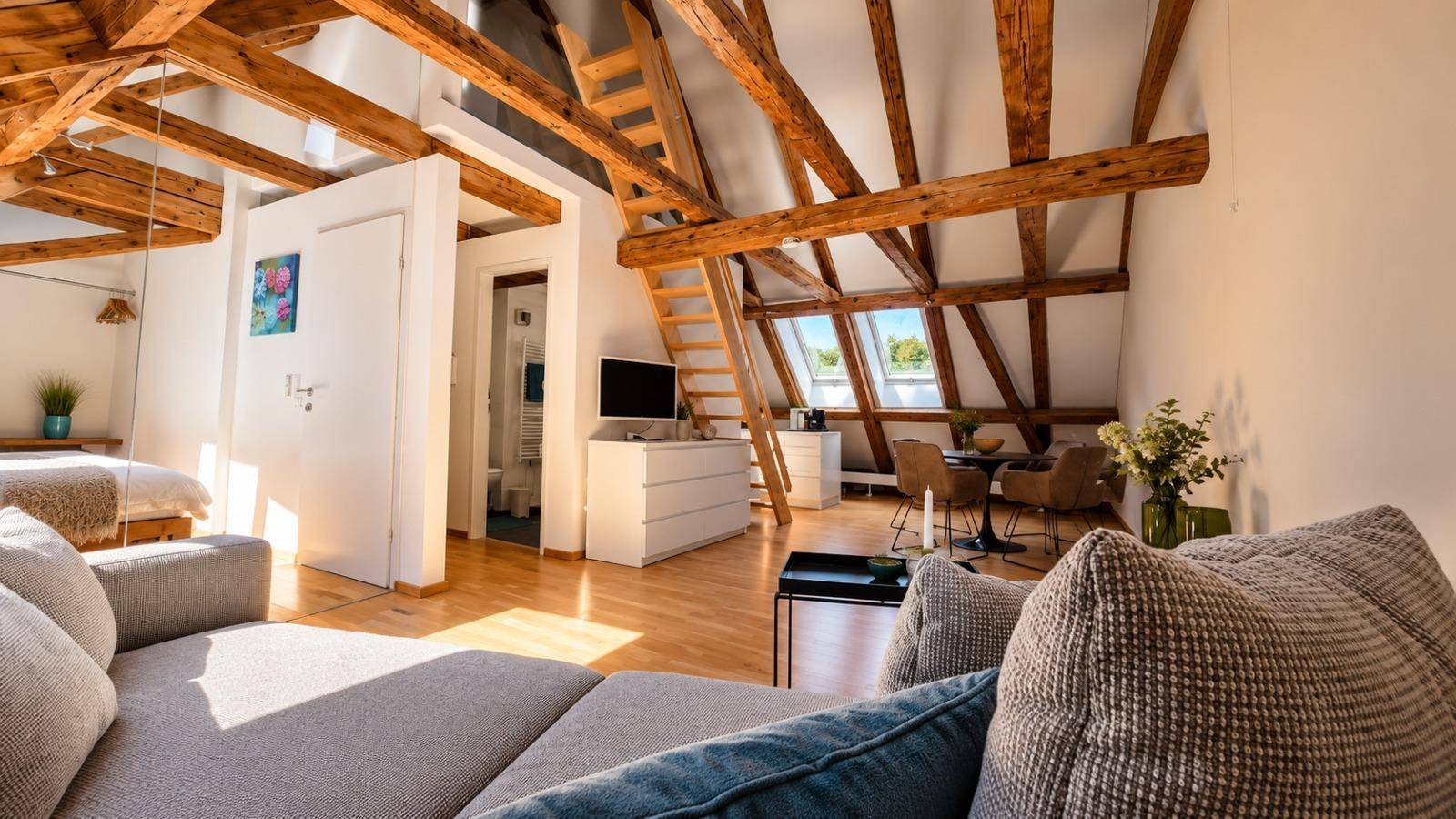 Geräumiger Wohnbereich im Dachgeschoss mit bequemer Couch, Holzbalken und Zugang zu einem Schlafbereich.